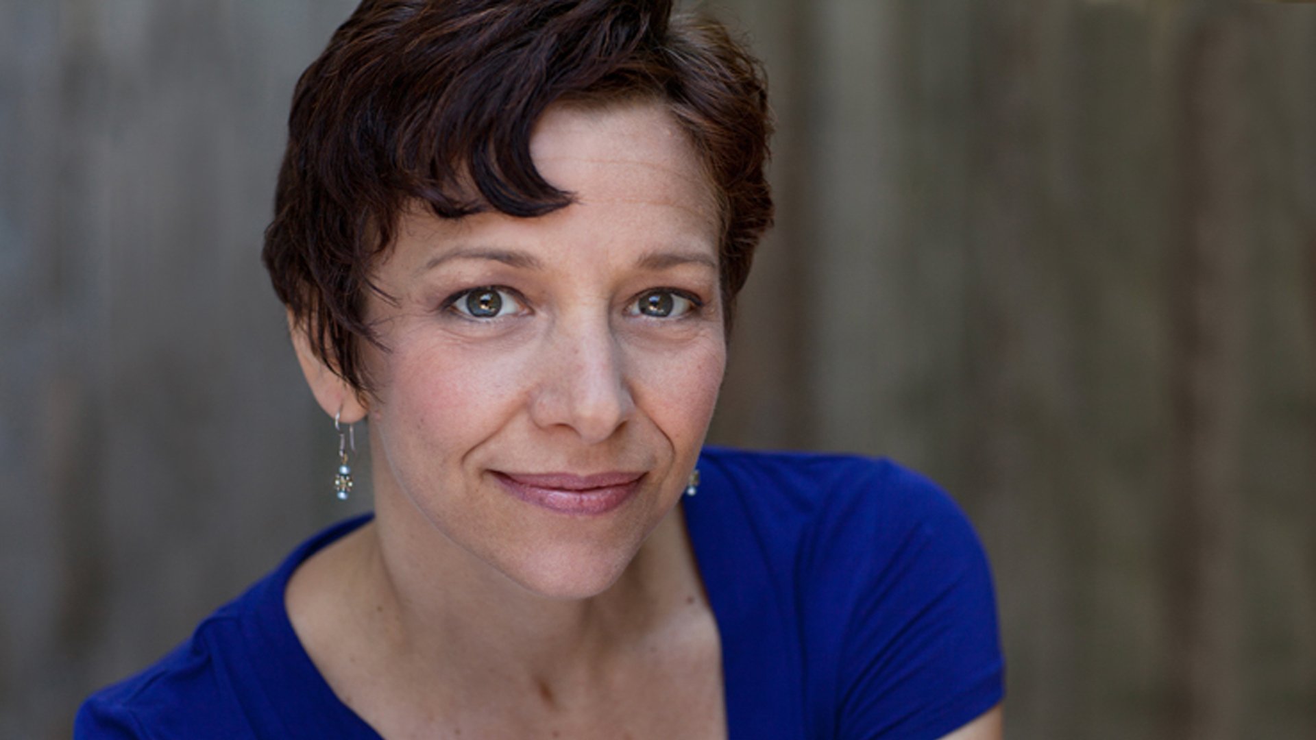 Close-up of a woman with short brown hair, wearing a blue top and earrings, smiling softly at the camera, with a blurred wooden background.