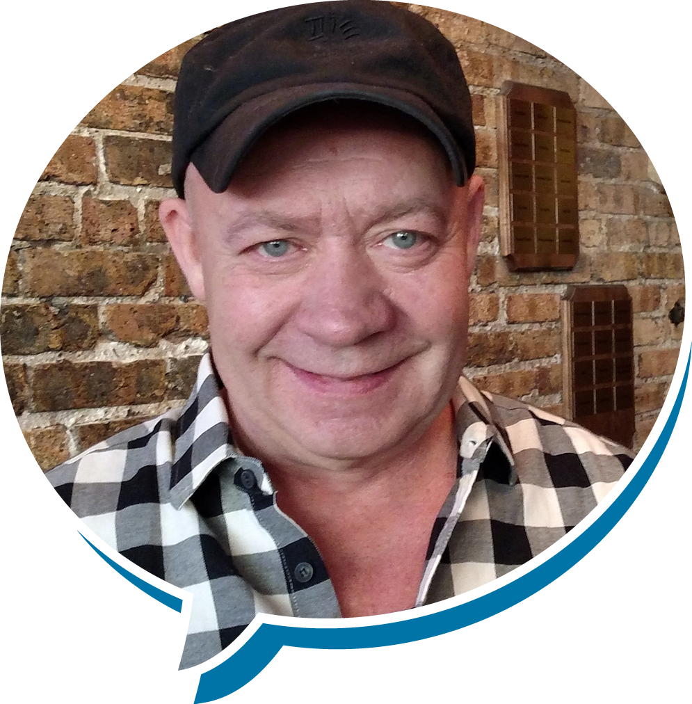 A smiling man with blue eyes, wearing a black cap and a black-and-white checkered shirt, standing in front of a brick wall.