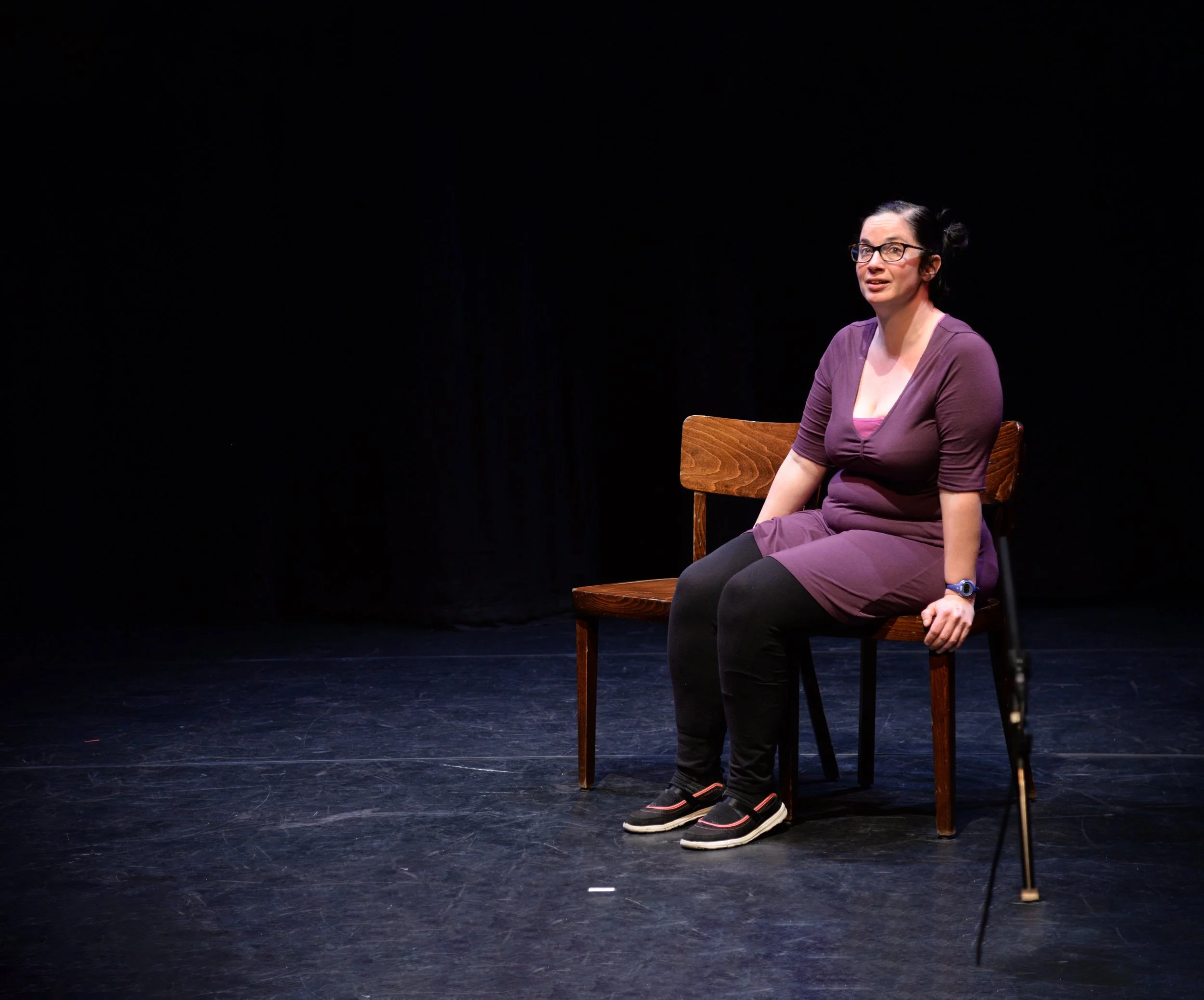 A woman with glasses and dark hair, seated on a wooden chair on a dark stage, wearing a purple dress and black leggings, with a cane beside her.