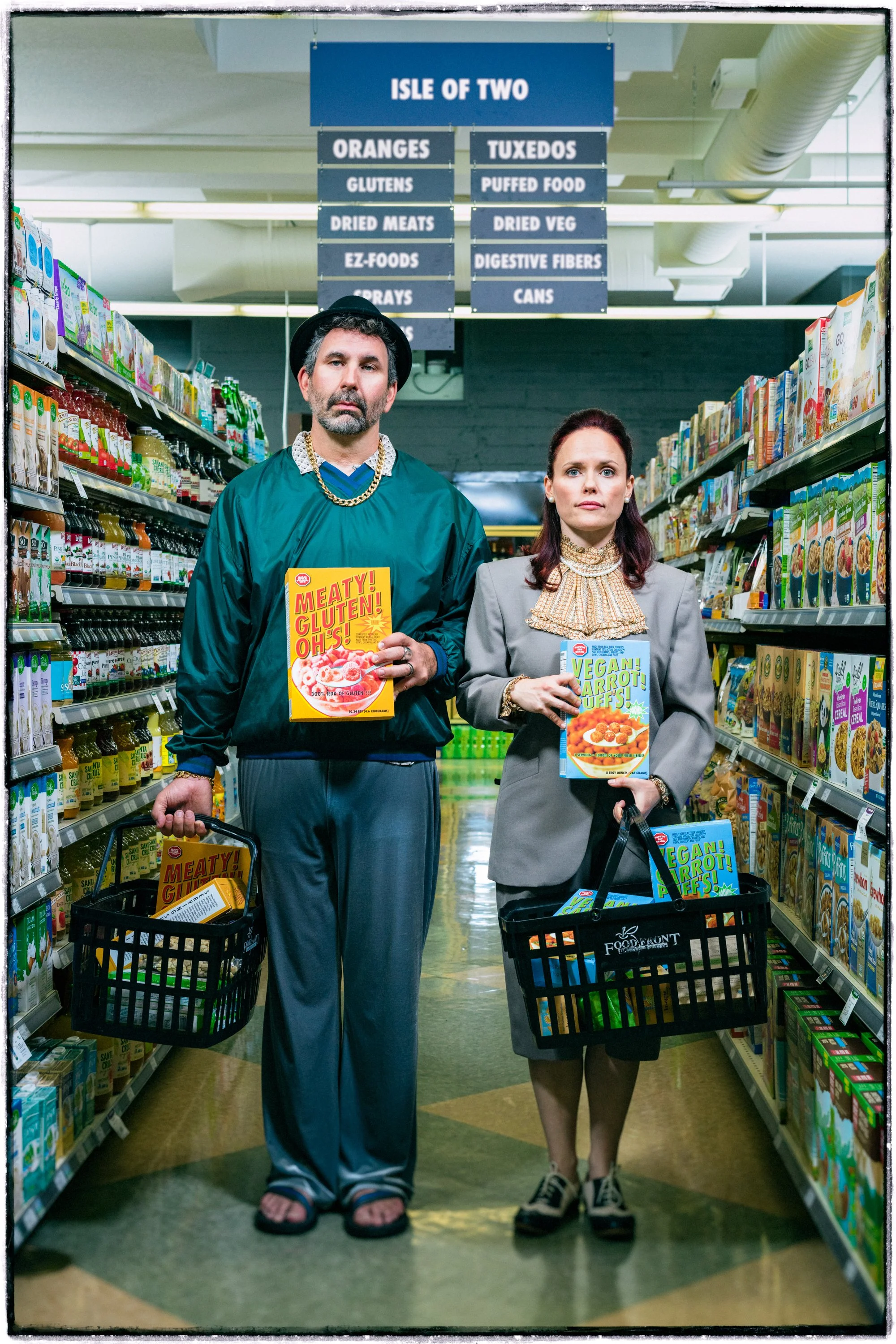 Two people standing in a grocery store aisle holding boxes of vegan carrot puffs and meatless gluten-free products, with a sign overhead listing aisle items like oranges, tuxedos, and canned foods.