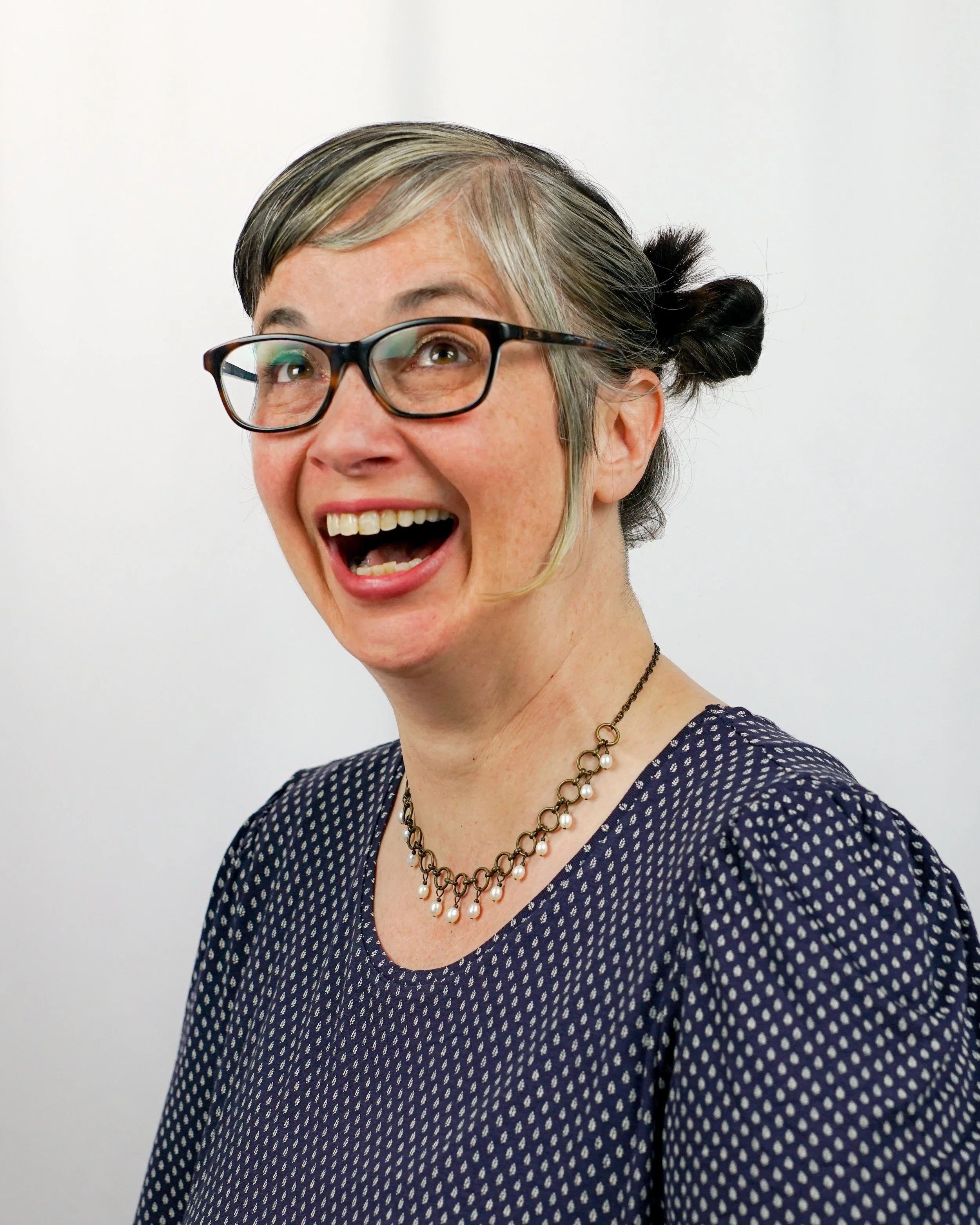 Woman with glasses smiling, wearing a navy blue patterned dress and a pearl necklace, against a plain white background.