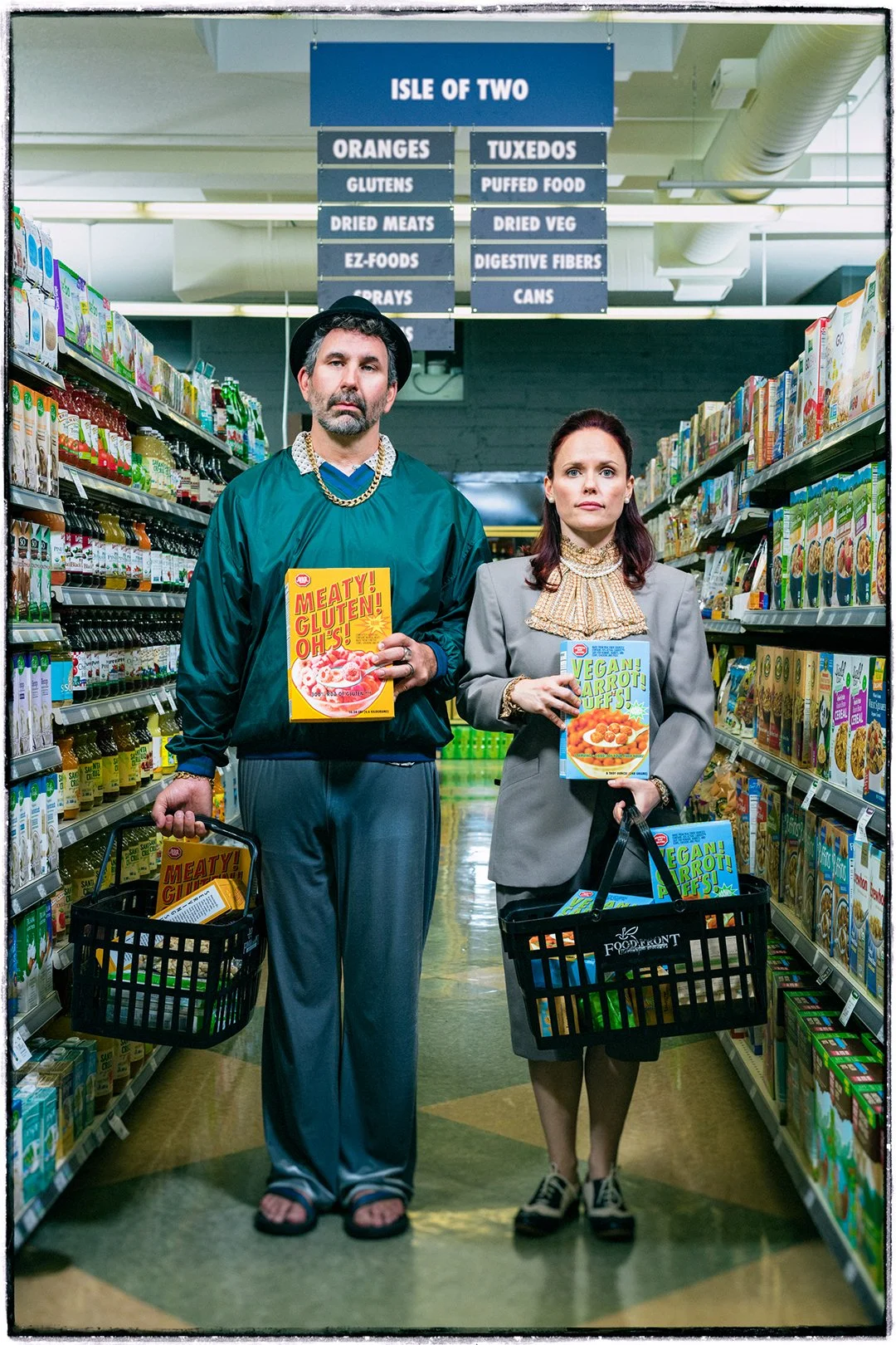 A man and woman standing in a grocery store aisle holding boxed products, with a sign above them that reads 'ISLE OF TWO.' The man is dressed casually with a hat and chain, holding a boxed item labeled 'Meaty! Gluten! Oh!.' The woman is dressed in business attire with a ruffled blouse, holding a box labeled 'Vegan! Carrot! Off!' Both are carrying shopping baskets with similar products.