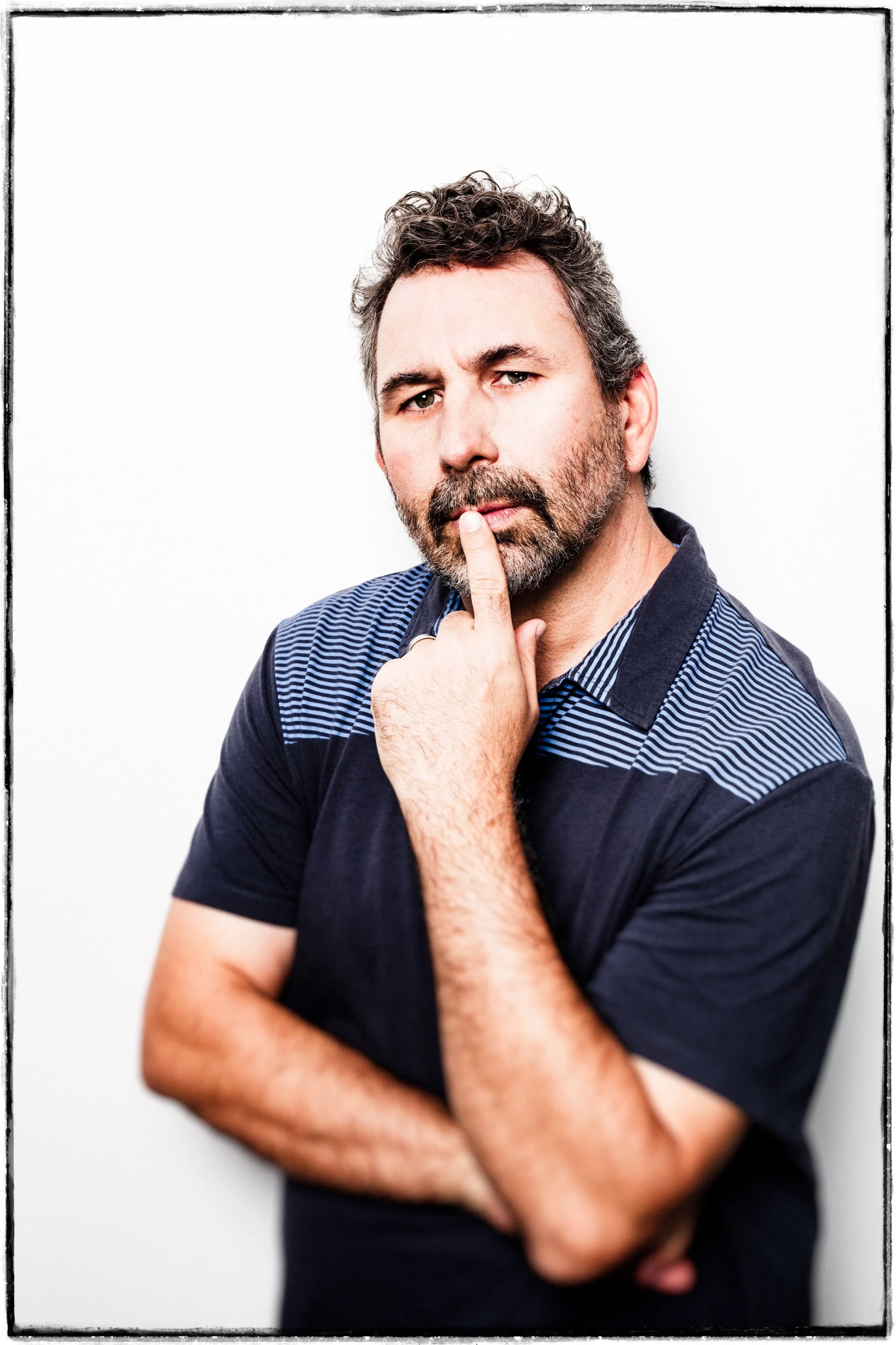 A man with dark hair and a beard, wearing a navy blue shirt with striped accents, posing thoughtfully with one finger on his lips against a white background.