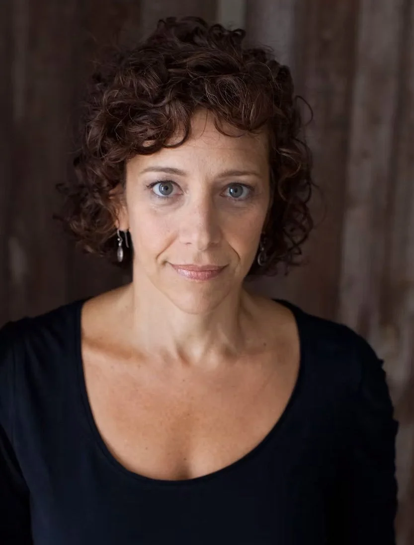 A woman with short, curly brown hair and blue eyes, wearing a black top and earrings, looking directly at the camera with a neutral expression against a wooden background.