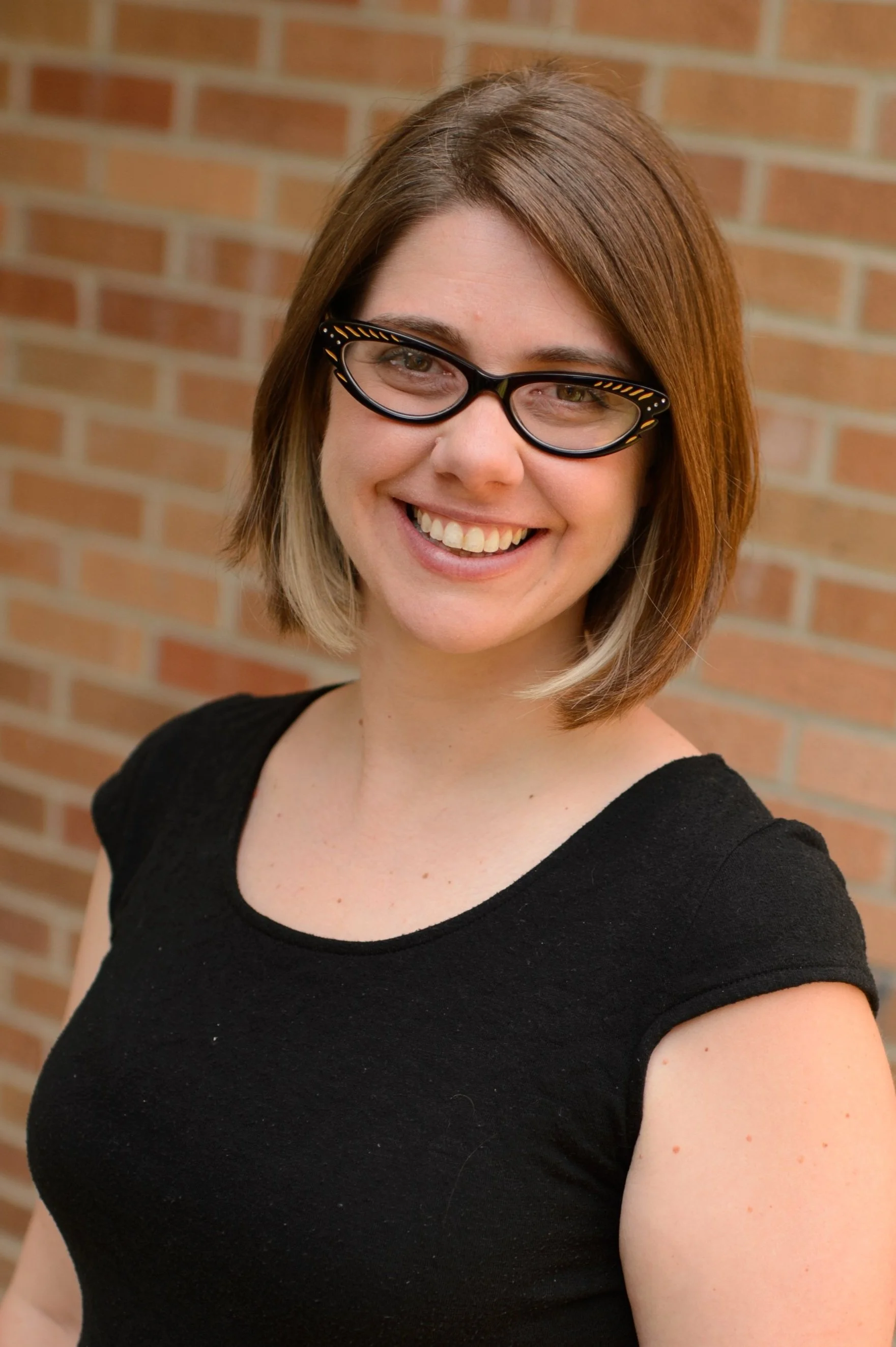 A woman with shoulder-length brown hair, wearing glasses and a black shirt, smiling, with a brick wall in the background.