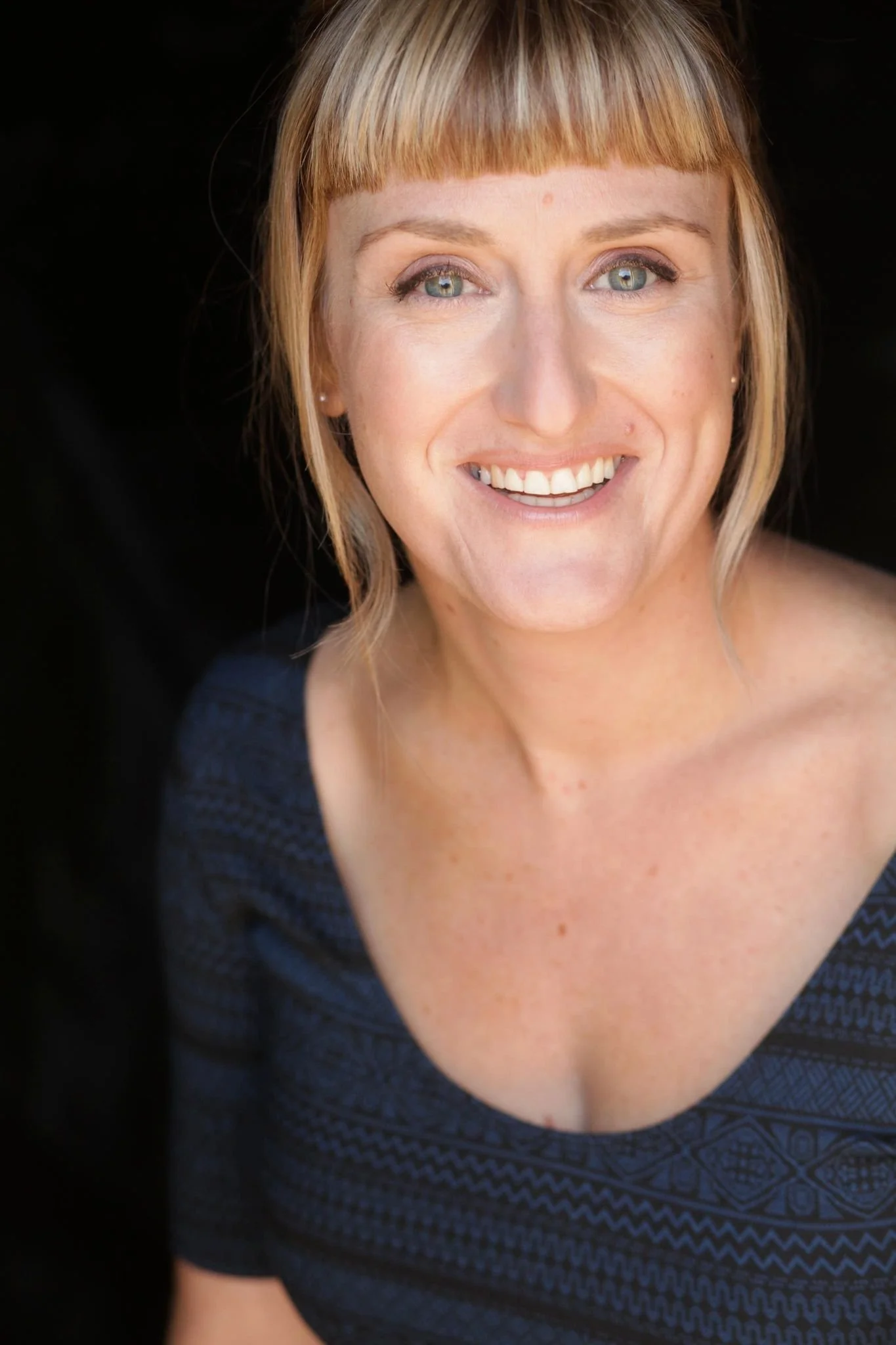 Close-up portrait of a smiling woman with blonde hair and light eyes, wearing a dark patterned top, against a black background.
