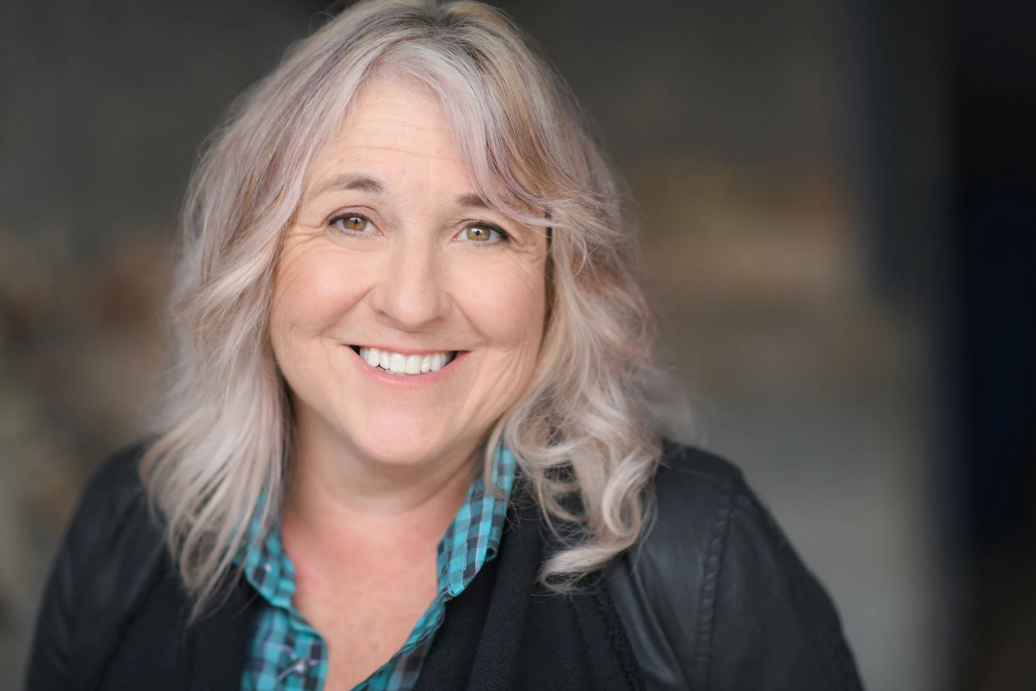 A woman with gray, wavy hair smiling outdoors, wearing a black jacket and a blue plaid shirt.