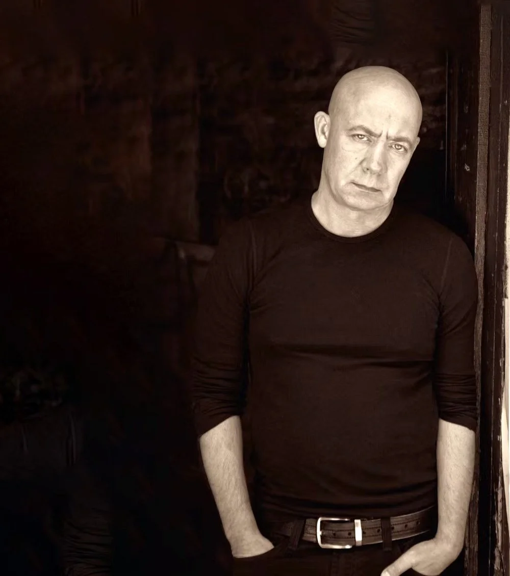 A bald man wearing a black long sleeve shirt standing against a dark background. He has a serious expression and his right hand is in his pocket.