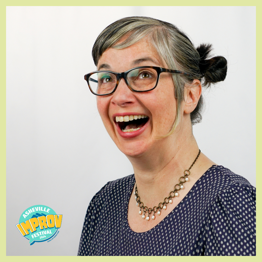 A smiling woman with glasses and gray hair styled in buns, wearing a polka dot blouse and a pearl necklace, at the Asheville Improv Festival 2024.