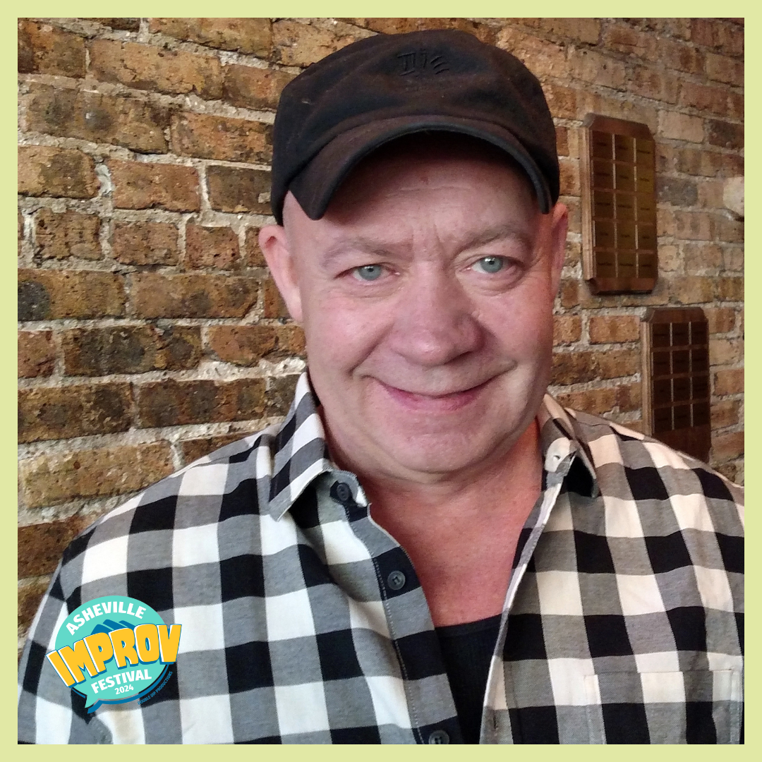 Smiling man wearing a black cap and black-and-white checkered shirt, standing in front of a brick wall with wooden decorations, at the Asheville Improv Festival 2024.