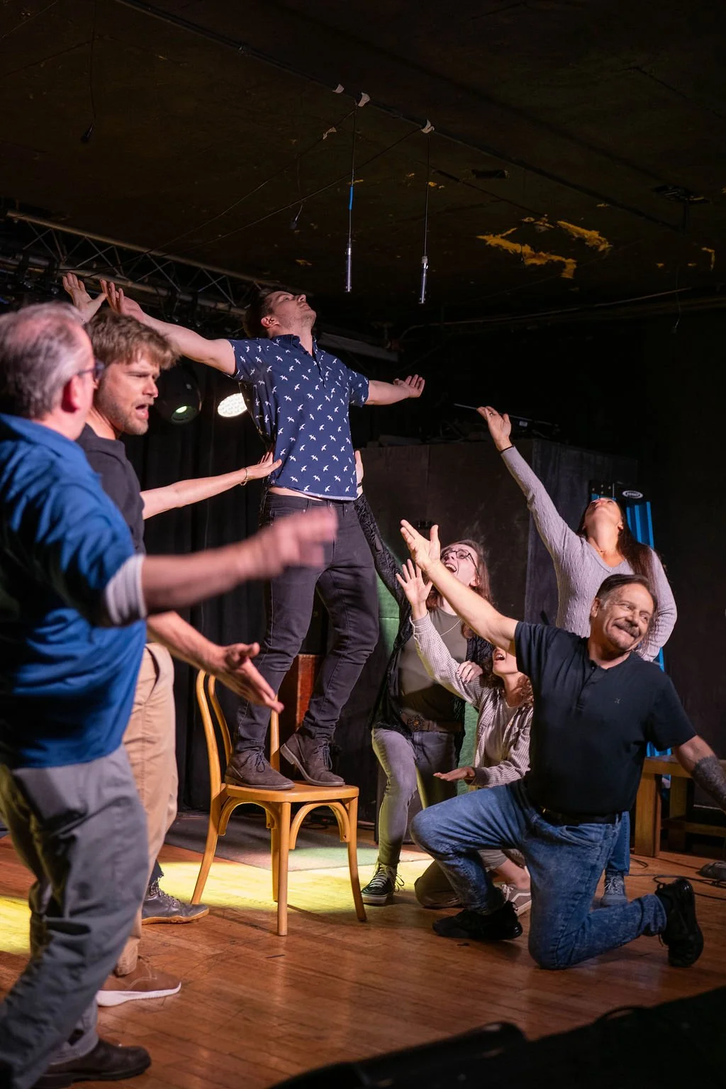 Group of people performing a theatrical act on a stage, with one person standing on a chair with arms outstretched, others surrounding and supporting him, expressive gestures, black backdrop, stage lighting, wooden floor.