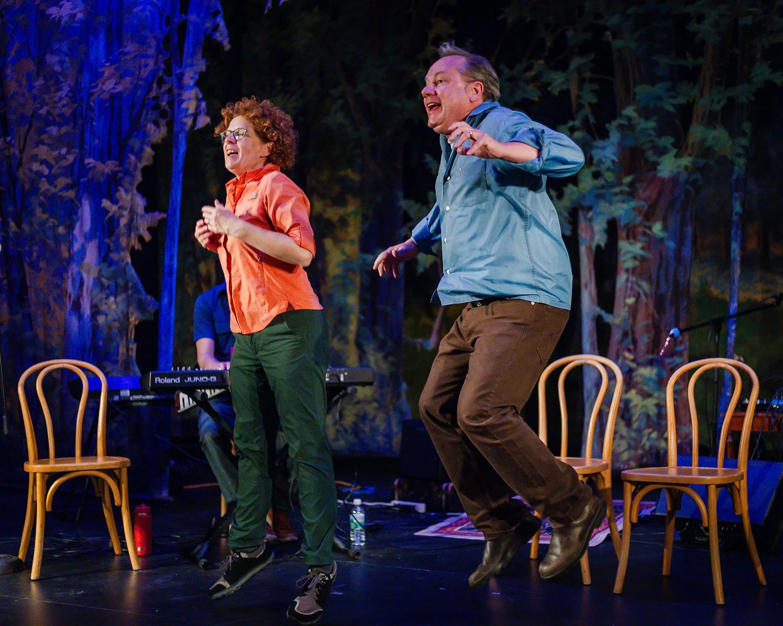 Two people dancing on stage with a forest background, a woman with curly hair wearing glasses, an orange jacket, and green pants, and a man with short hair in a blue jacket and brown pants, with chairs and musical equipment visible.
