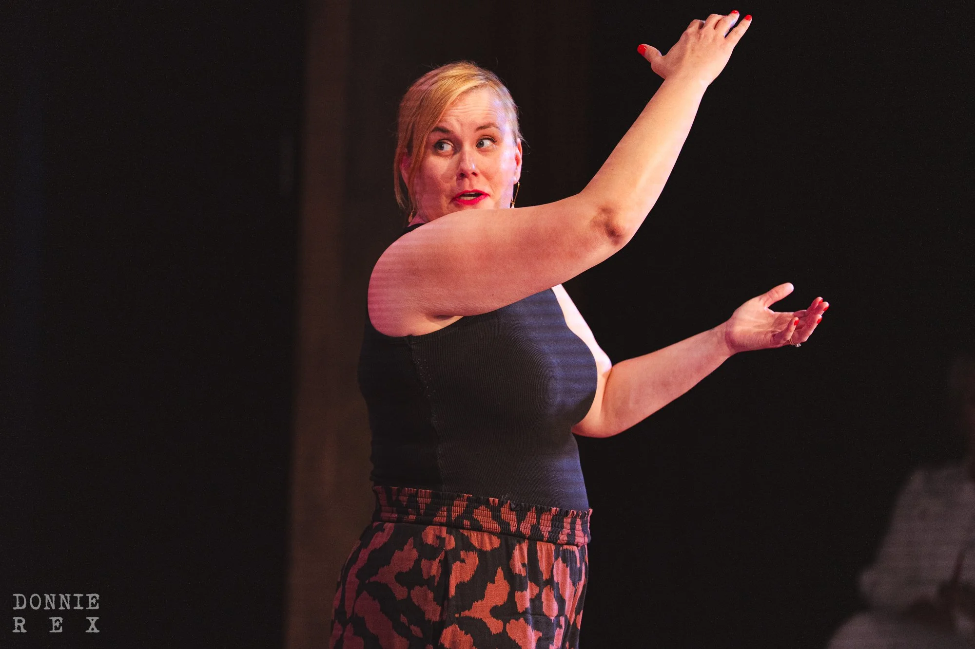 A woman with blonde hair, wearing a black sleeveless top and red leopard print pants, is performing on stage with her arms raised.