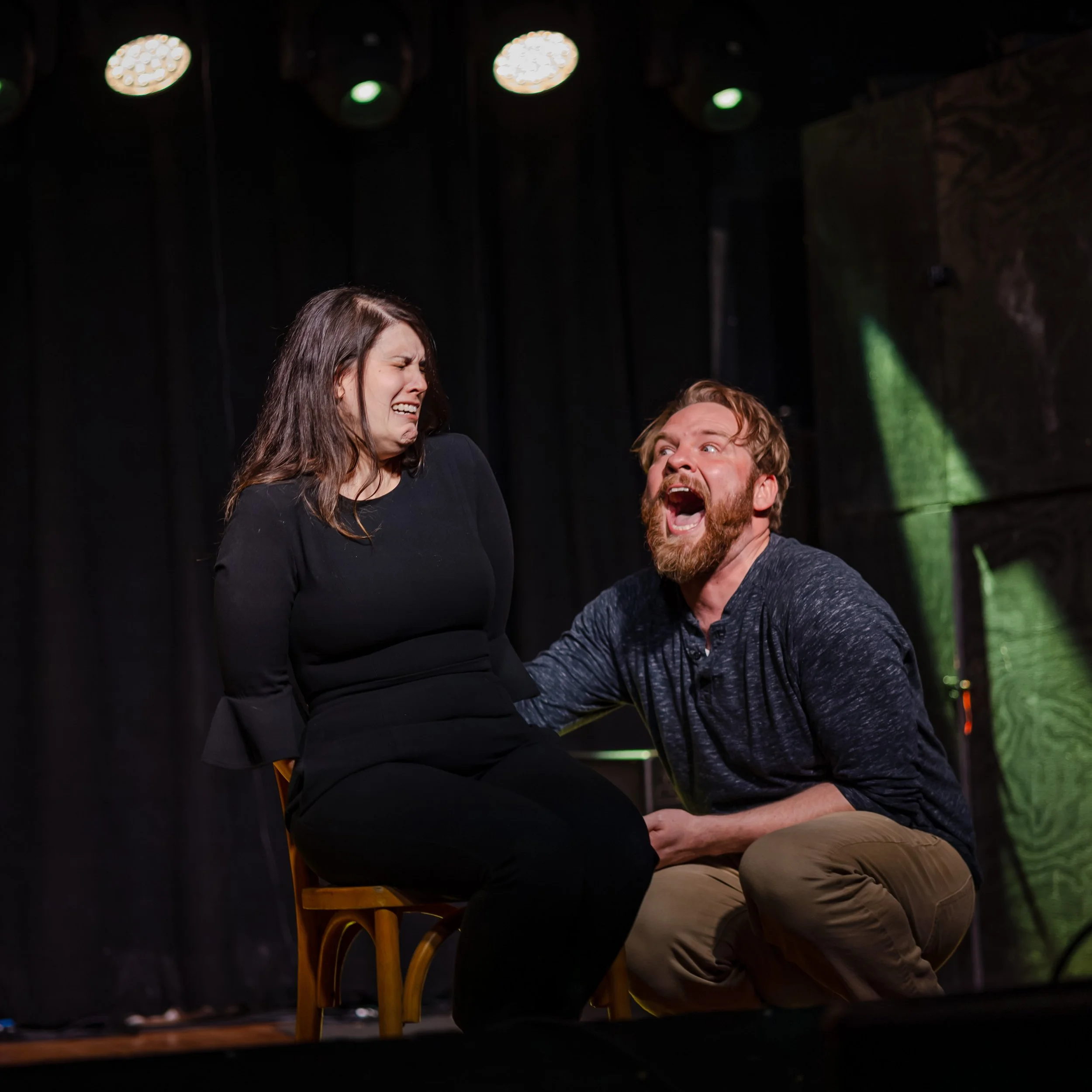 A woman sitting on a chair with her eyes closed and an expression of discomfort, while a man kneeling beside her, appears to be shouting or crying out, with both on a stage with black curtains and overhead stage lights.