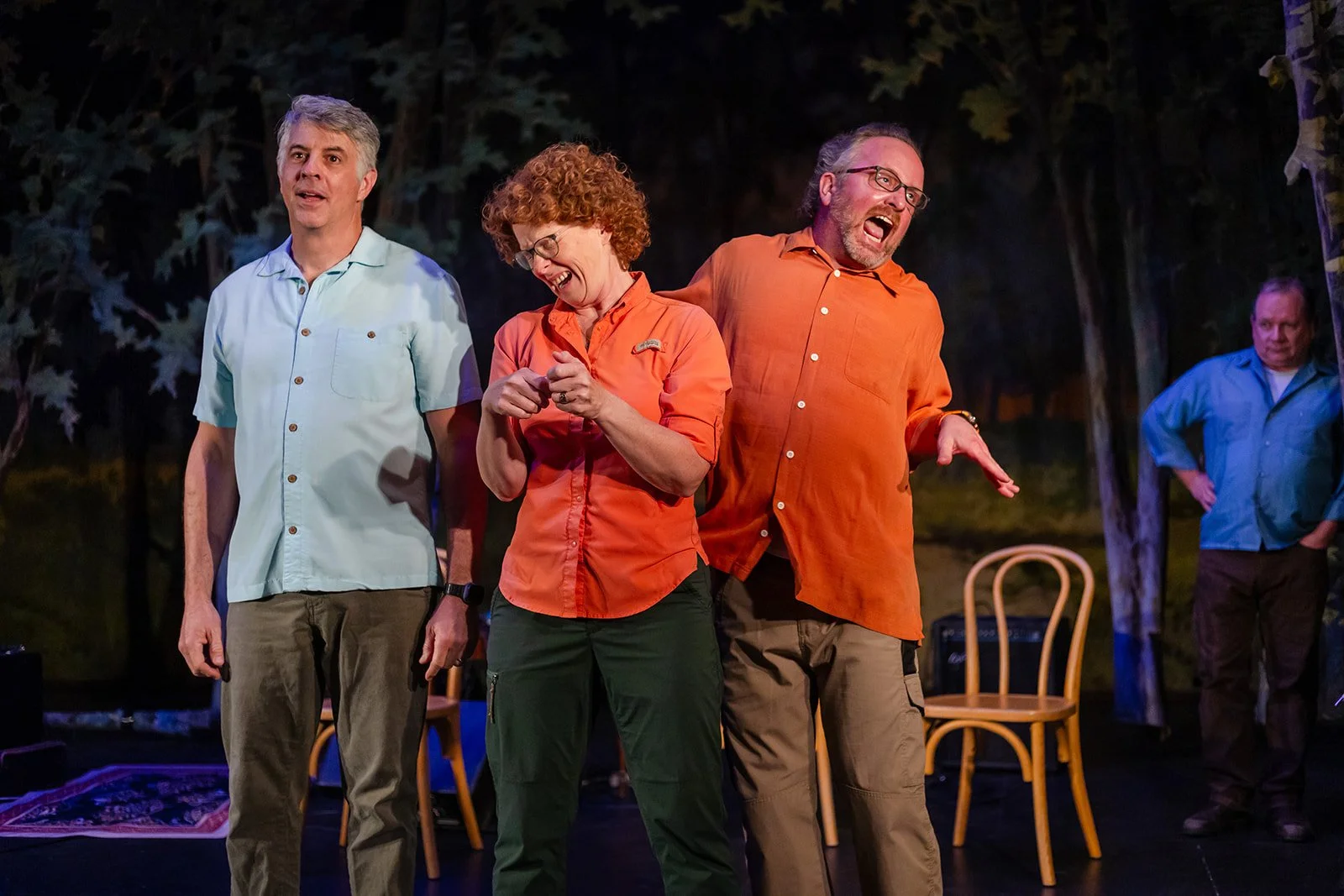 Four people on stage during a performance, with two men and one woman in the foreground, all wearing casual shirts, in front of chairs and trees.