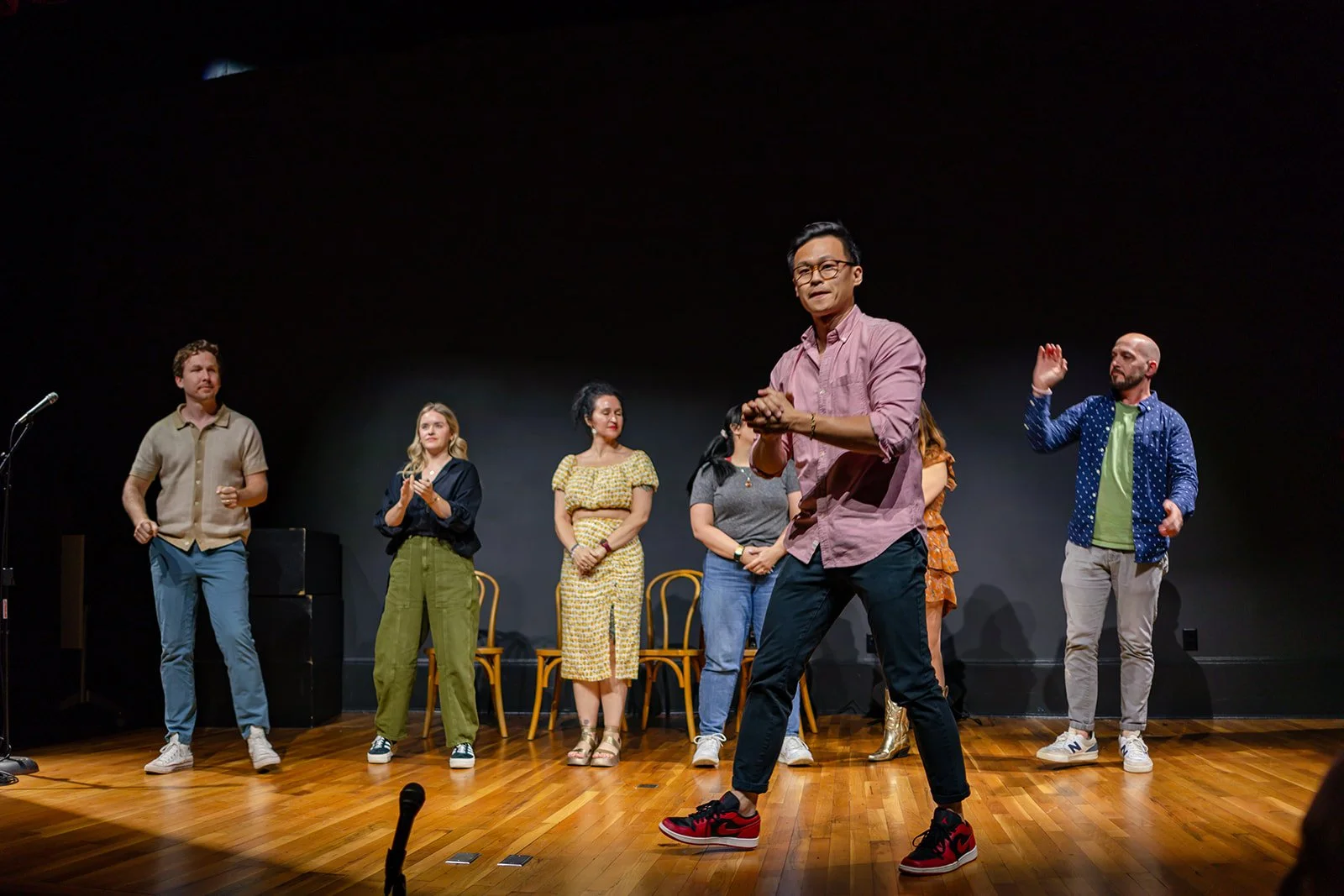 Group of people on stage, some clapping, with one man dancing in front, in a black room with a wooden floor.