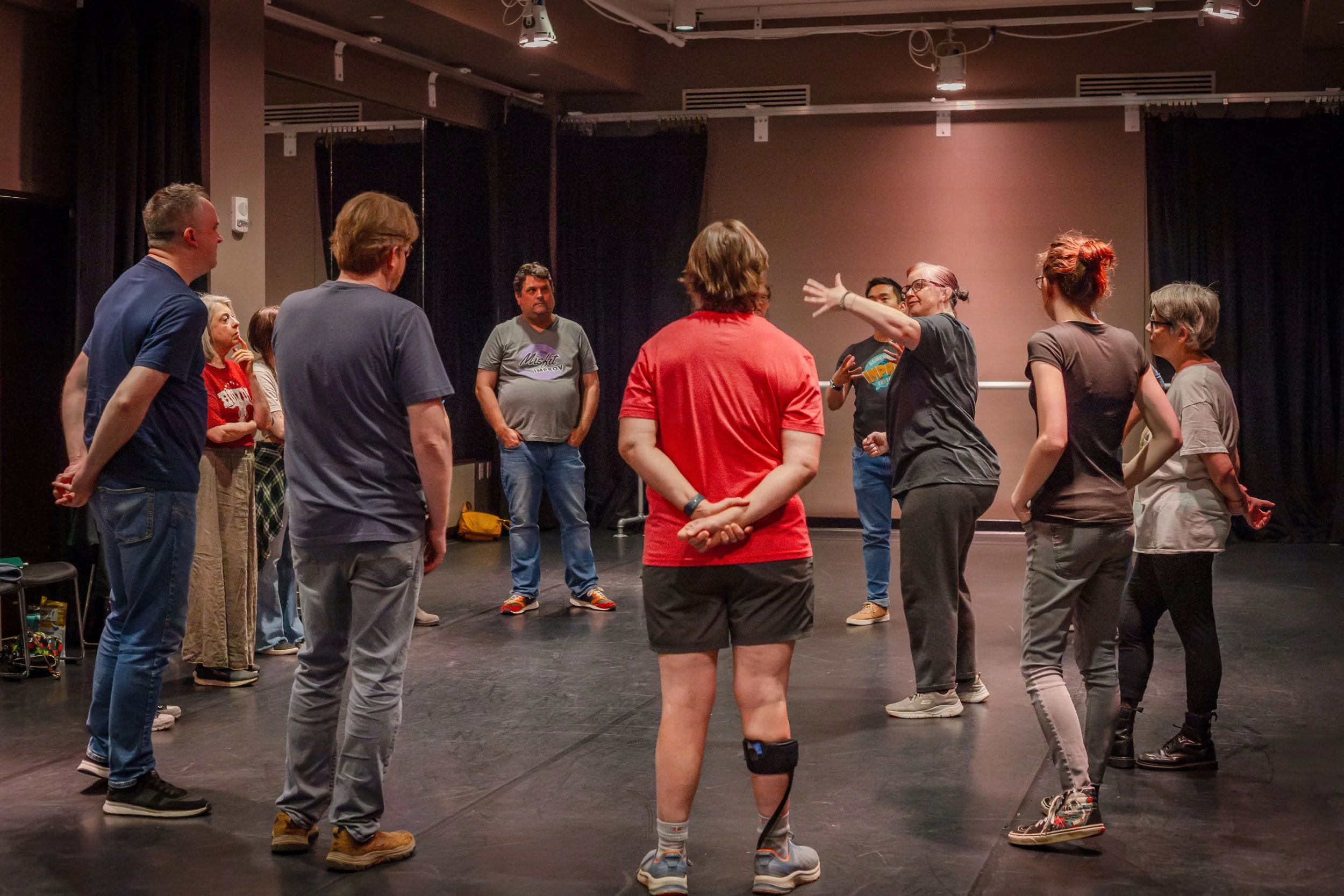 A group of people in casual clothing gathered in a circle in a theater or rehearsal space, with one woman speaking or leading the discussion.