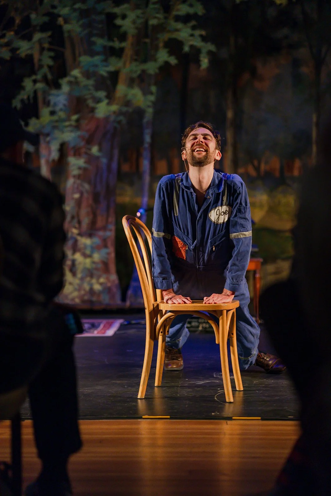 A man in a blue jumpsuit with a name tag that says 'Joe' is kneeling on a chair with his hands on the seat, smiling and laughing during a theatrical performance set in a forest backdrop.