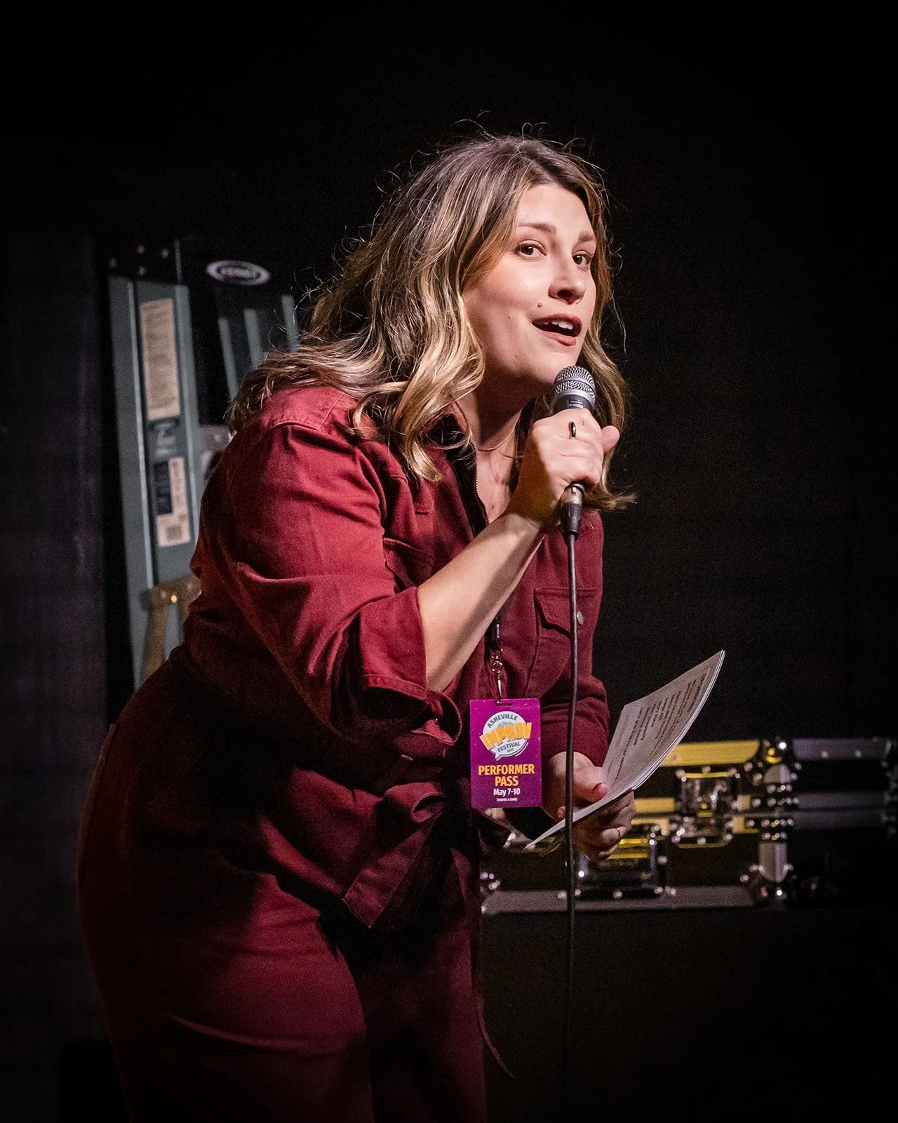 A woman with wavy blonde hair wearing a maroon outfit stands on stage, holding a microphone and a paper, speaking or singing at a performance event.