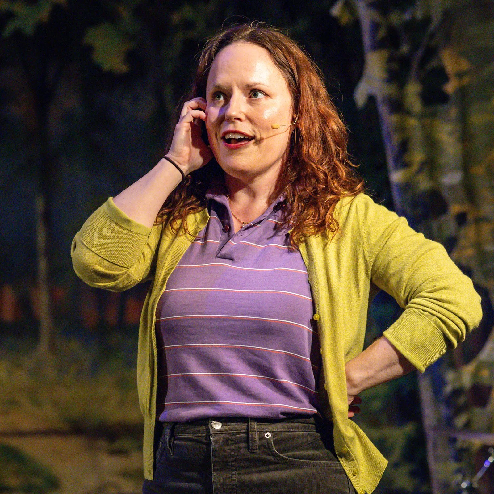 A woman with curly red hair speaking on a headset microphone in an outdoor setting, wearing a purple striped polo shirt and a yellow cardigan.