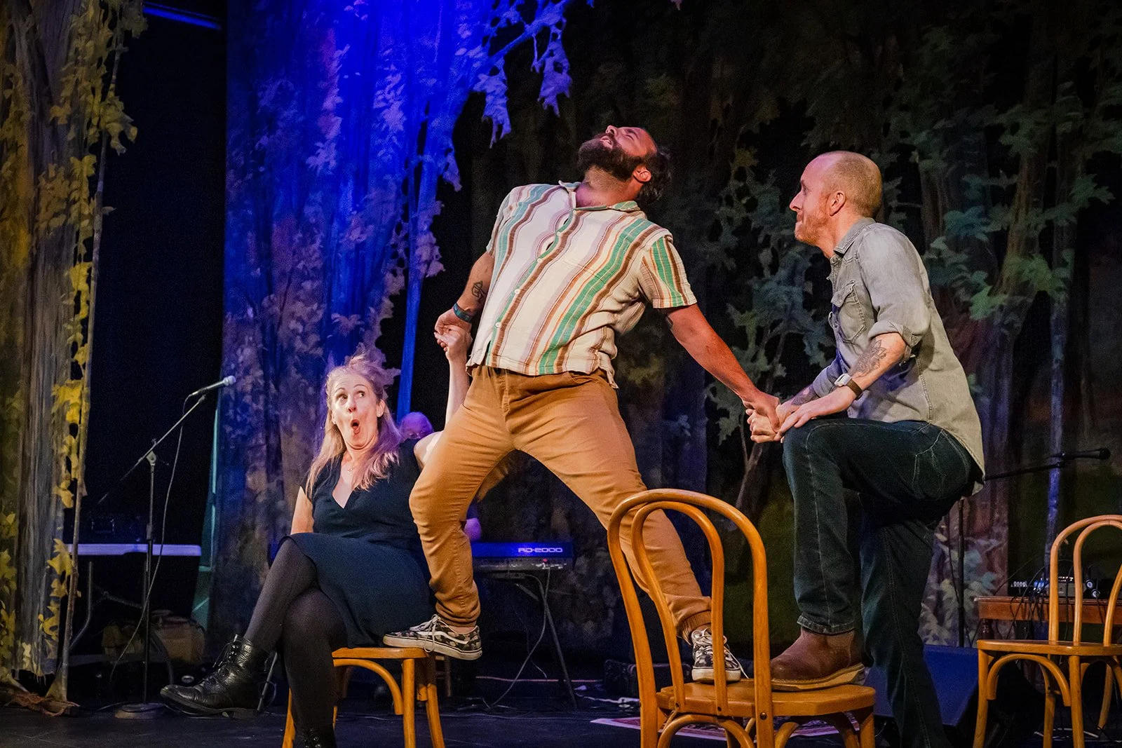 Three people on stage during a theatrical performance with one sitting on a chair, one standing on a chair, and another kneeling on a chair. The woman seated appears surprised, while the two men are holding hands and engaged in an intense scene. The stage has a backdrop of trees.
