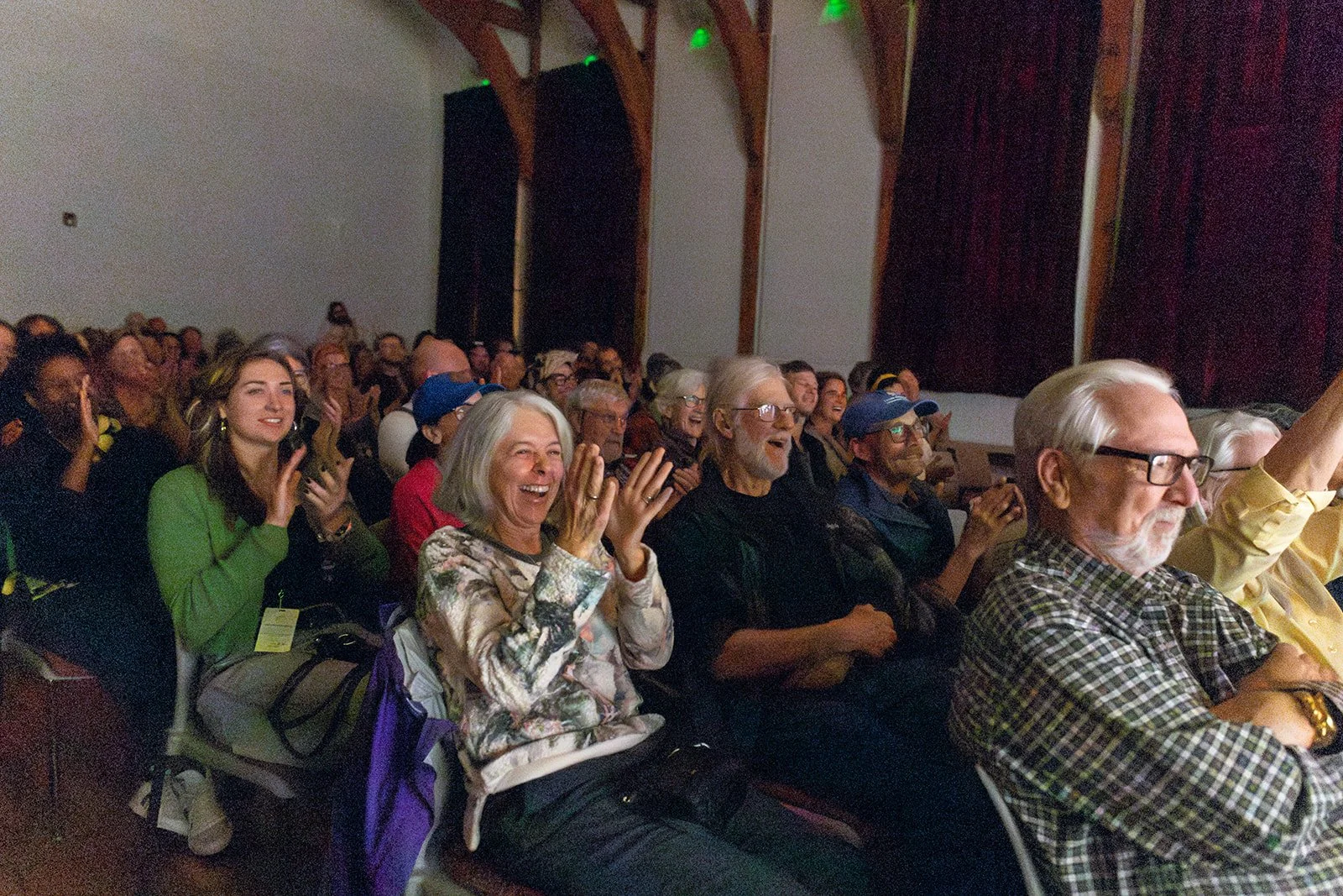 An audience watching a performance, smiling and laughing in a large room with wooden beams and dark red curtains.