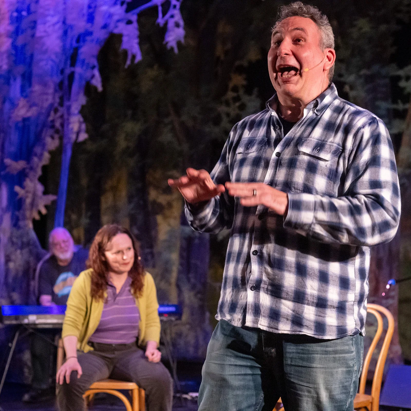 Man in a plaid shirt speaking on stage with two women in the background, outdoor setting with trees and purple lighting.