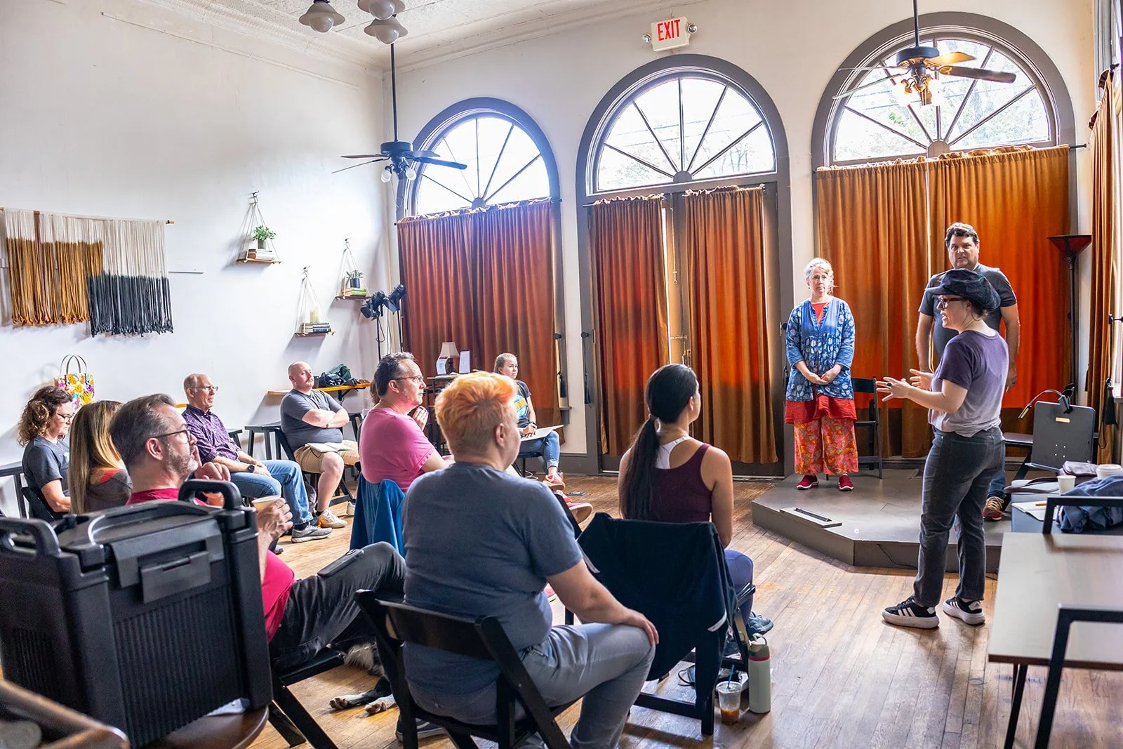 A group of people sitting and listening to two women speaking on a small stage in a brightly lit room with orange curtains, high arched windows, and ceiling fans.