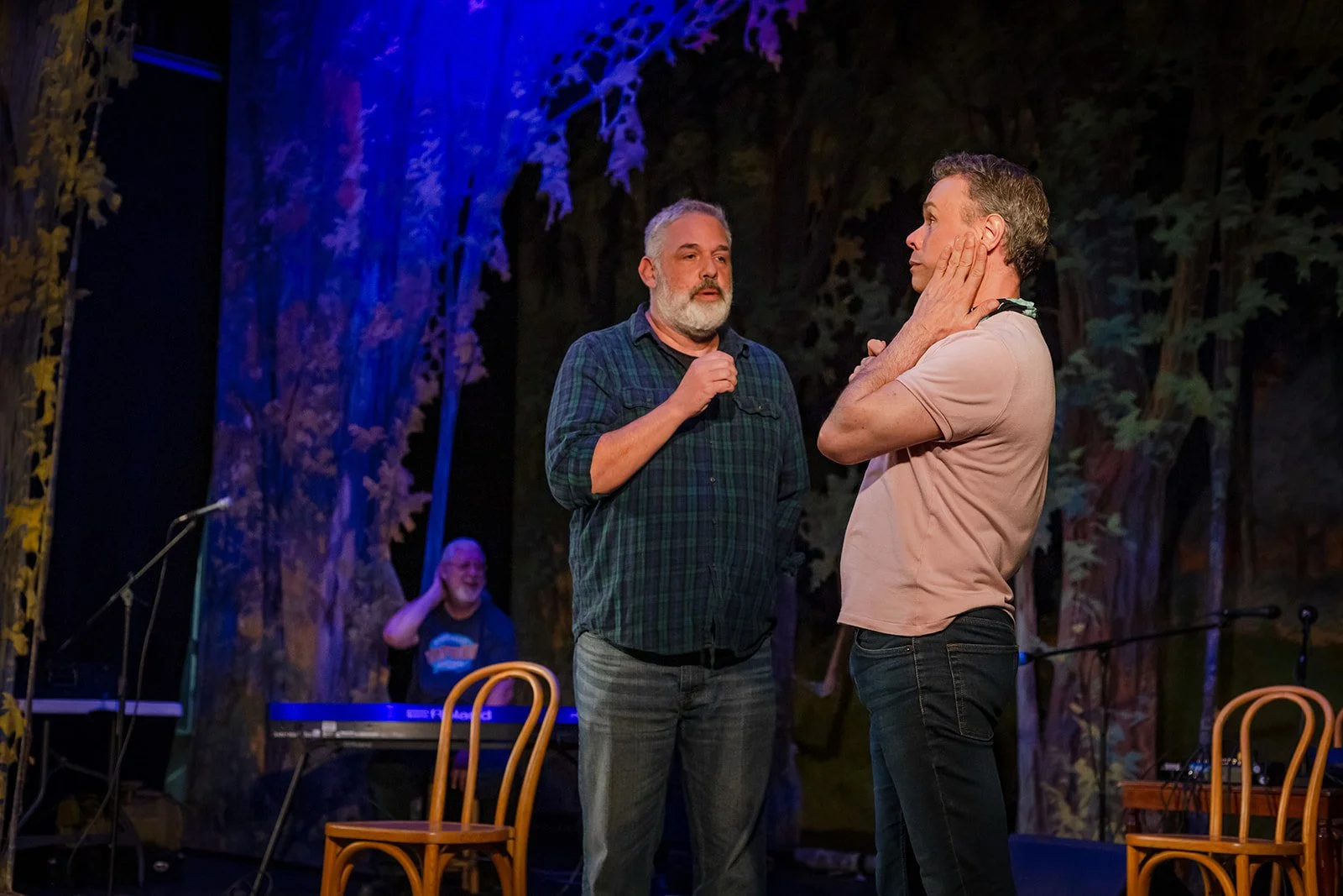 Two men are on stage in a theatrical play, with one man touching the other's face as they converse. A woman playing a keyboard is laughing in the background.