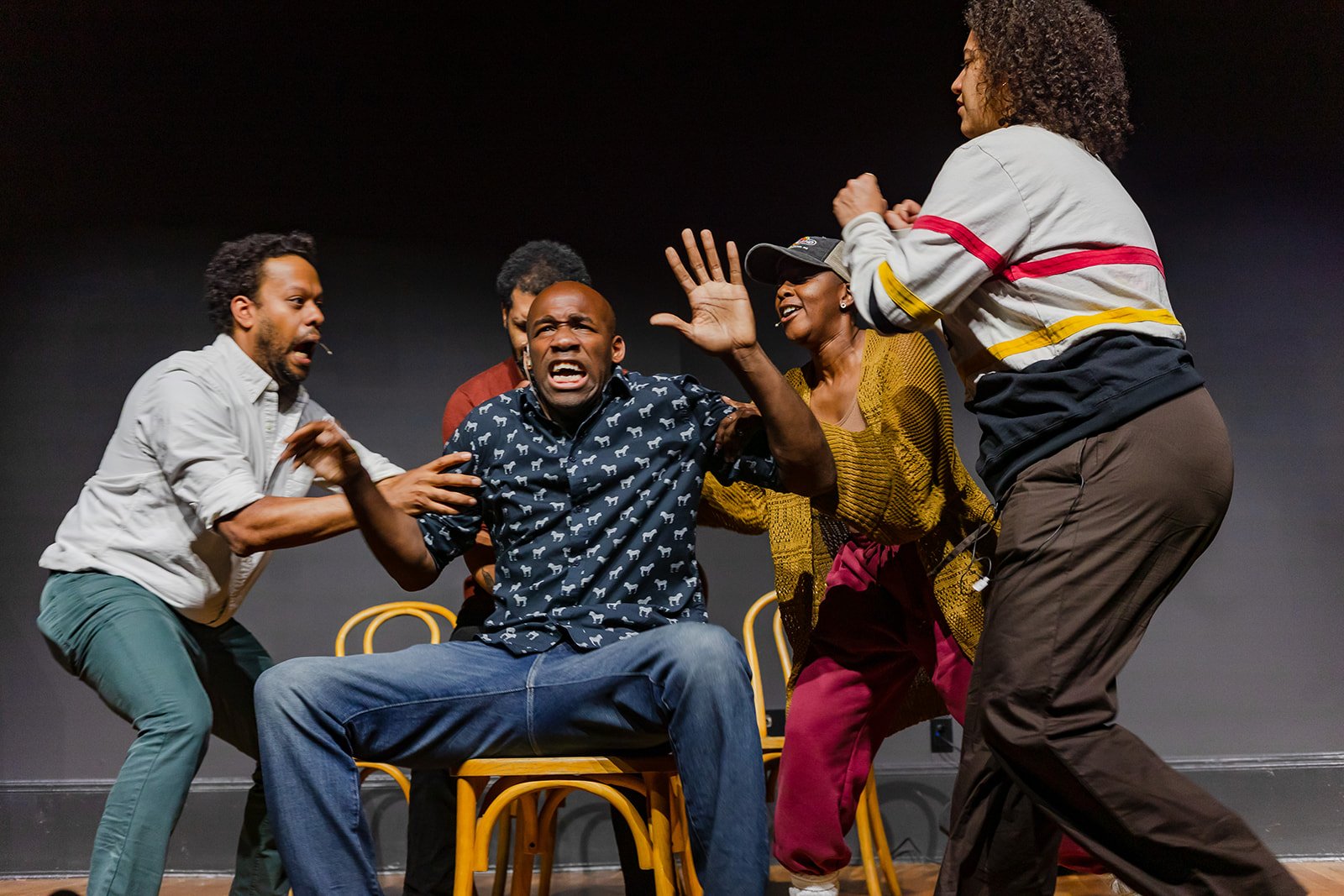 Group of people on stage engaged in an intense dramatic scene, with a man seated in the center expressing anger or fear, surrounded by others reacting emotionally.