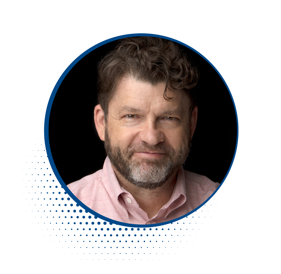 Close-up portrait of a middle-aged man with curly brown hair and a beard, wearing a pink collared shirt against a black background.