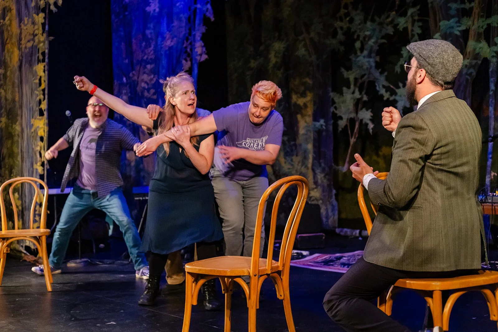 People staged in a theatrical scene, with a woman in black dress being restrained by two women, and three men in the background, set against a forest-themed backdrop.