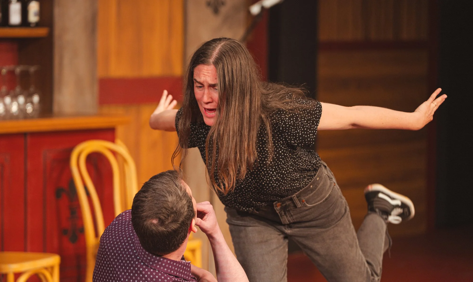 A woman is yelling at a man during a theatrical performance, with her arms extended and a distressed expression.