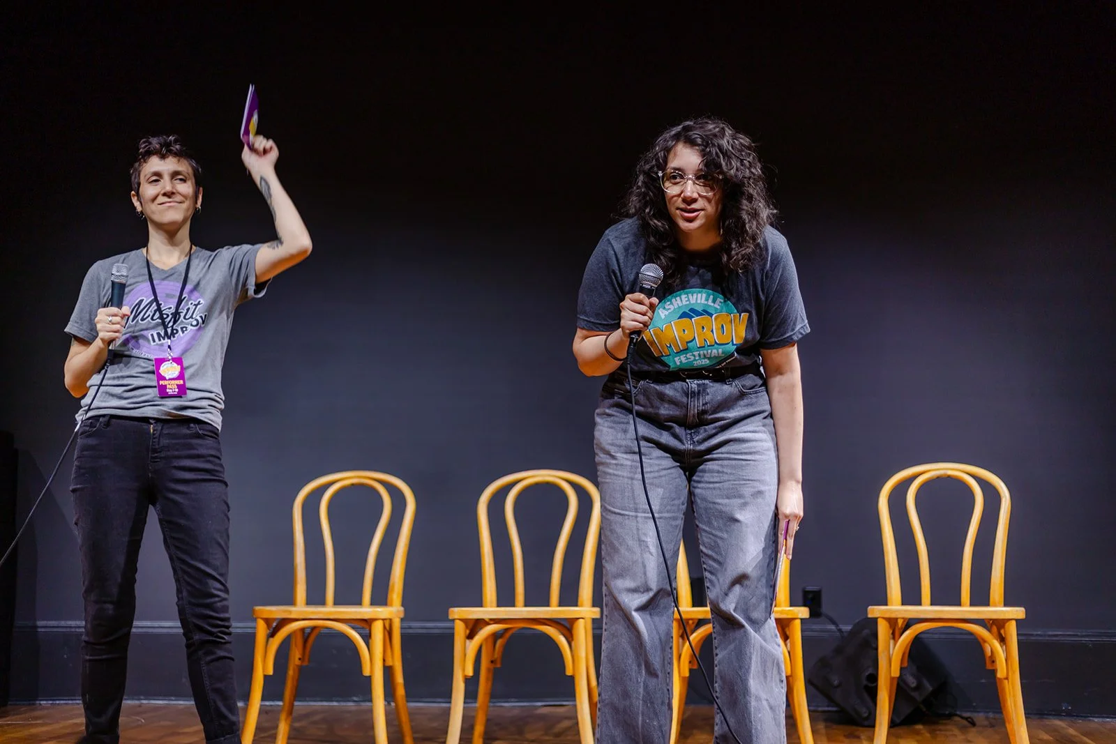 Two women performing stand-up comedy on stage with three empty chairs behind them, holding microphones.