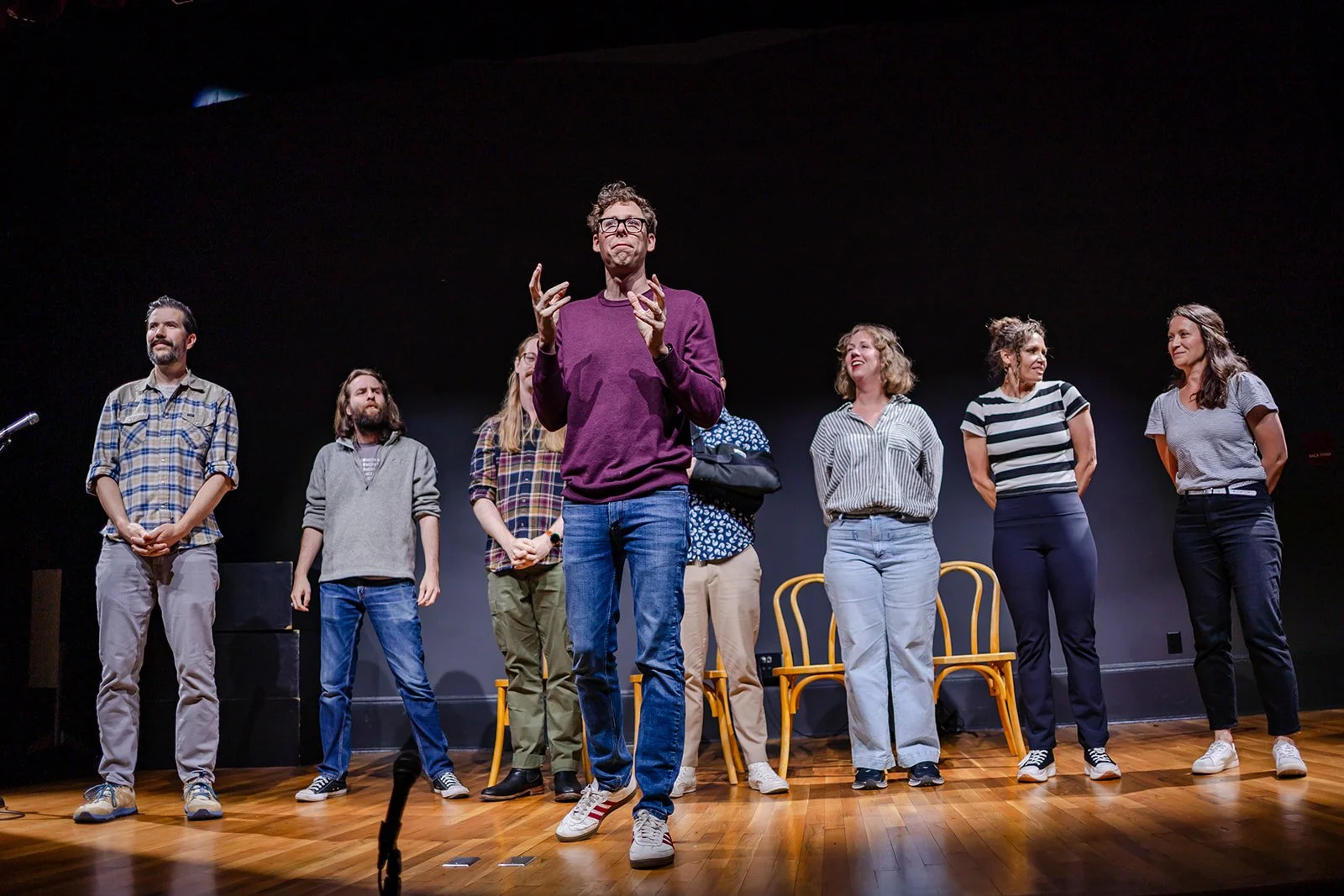 A group of eight people standing on a stage, with one person in the center speaking or performing, and the rest watching or waiting. The stage has a black background and wooden floor, with yellow chairs behind the group.