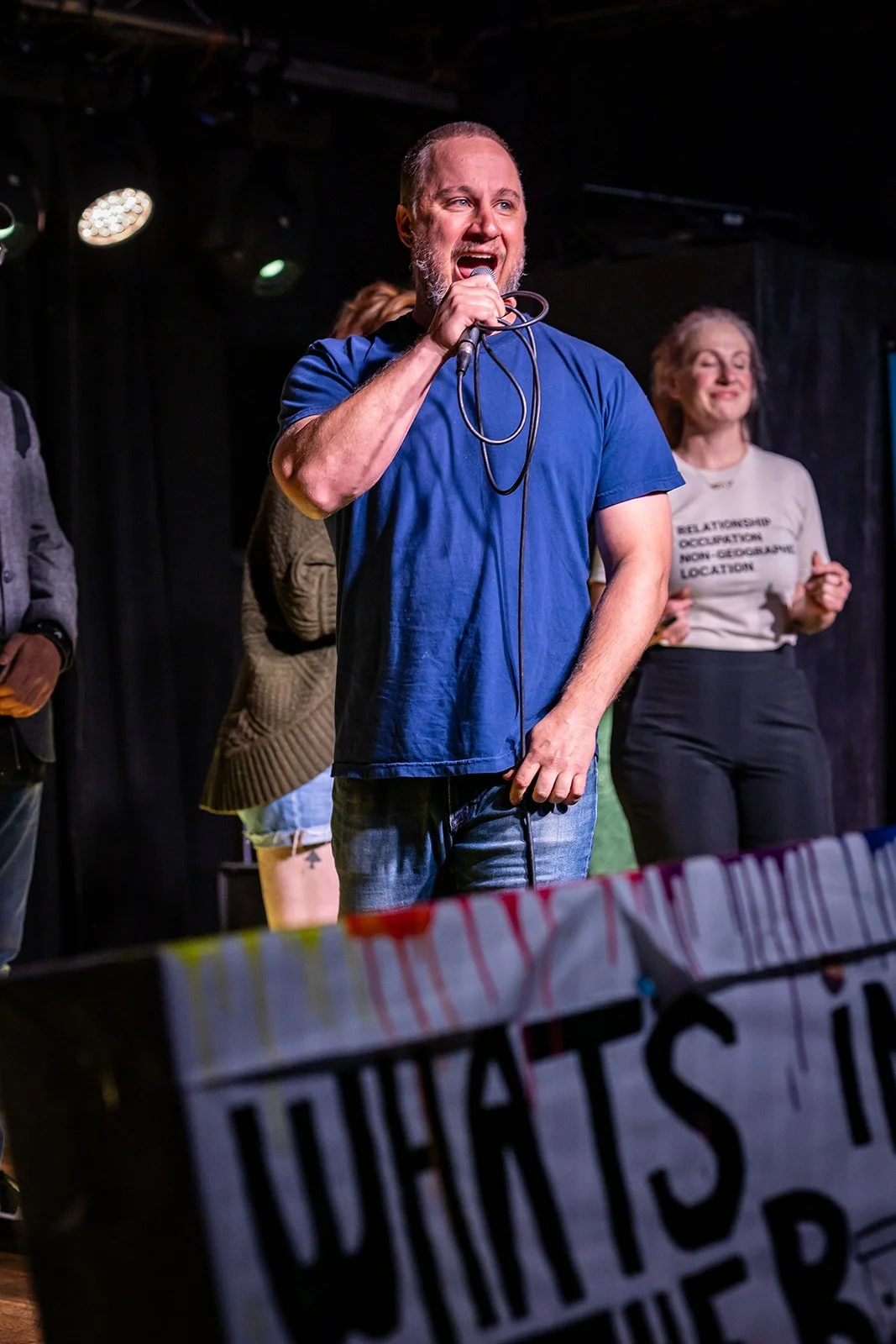 A man with a beard wearing a blue t-shirt speaking into a microphone on stage, with two women behind him, one in a gray sweater and the other in a white shirt, at a public event or protest.