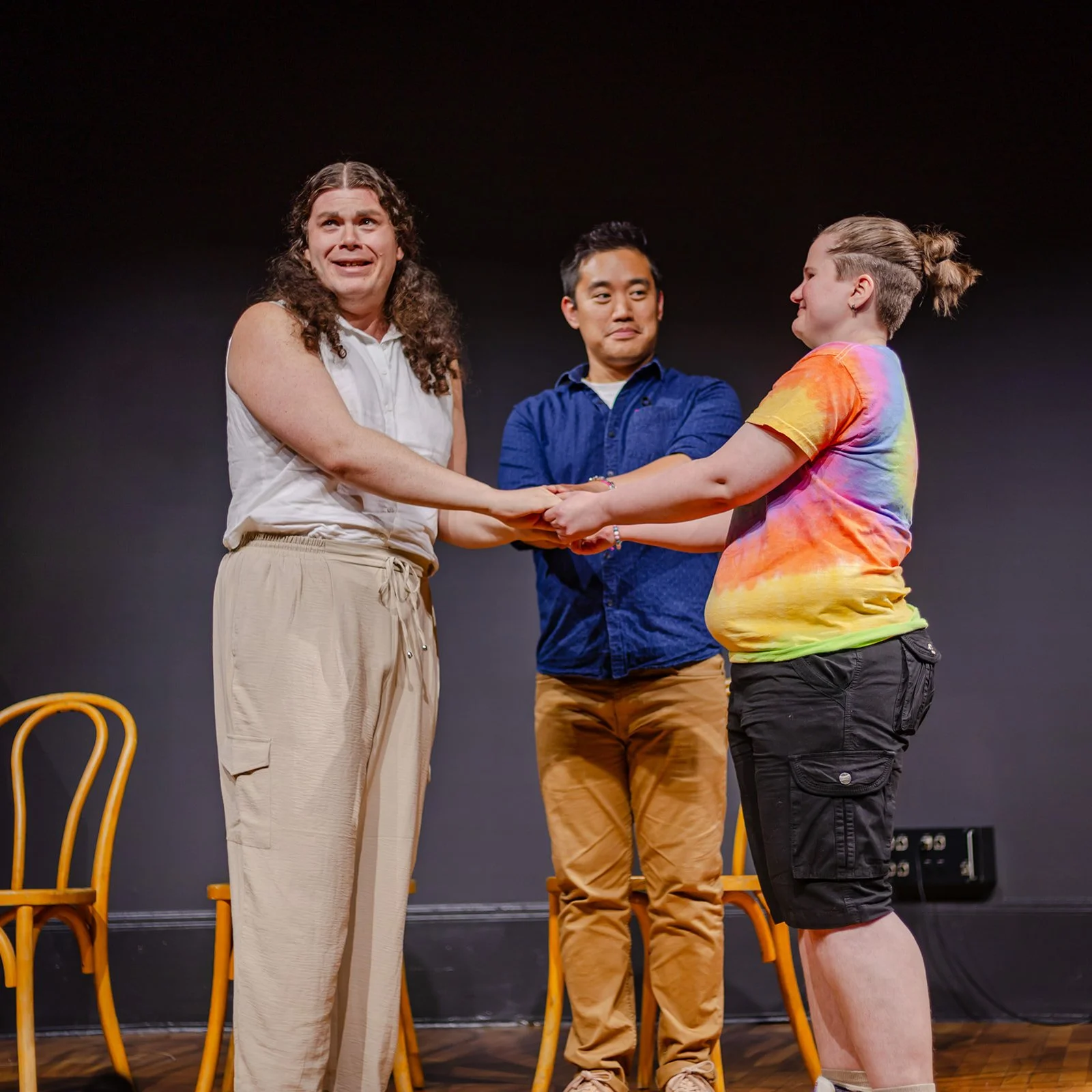 Three people on a stage holding hands, engaging in a group activity or performance, with chairs in the background.