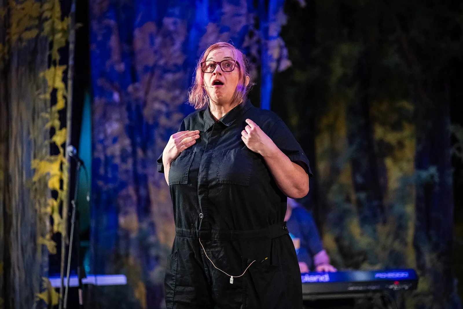 A woman with glasses and pink hair on a stage, wearing a black jumpsuit, appears to be in the middle of speaking or performing, with a forest-themed set and a keyboard in the background.
