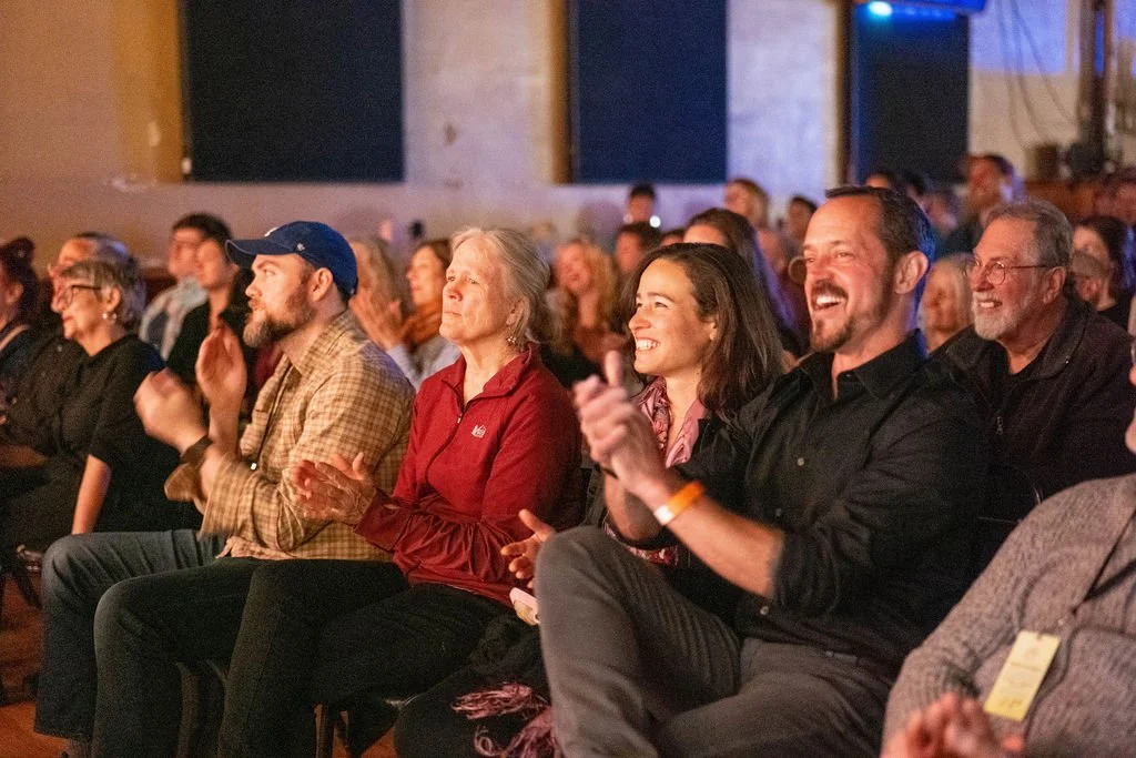 Audience members sitting in a theater, clapping and smiling during a live event.