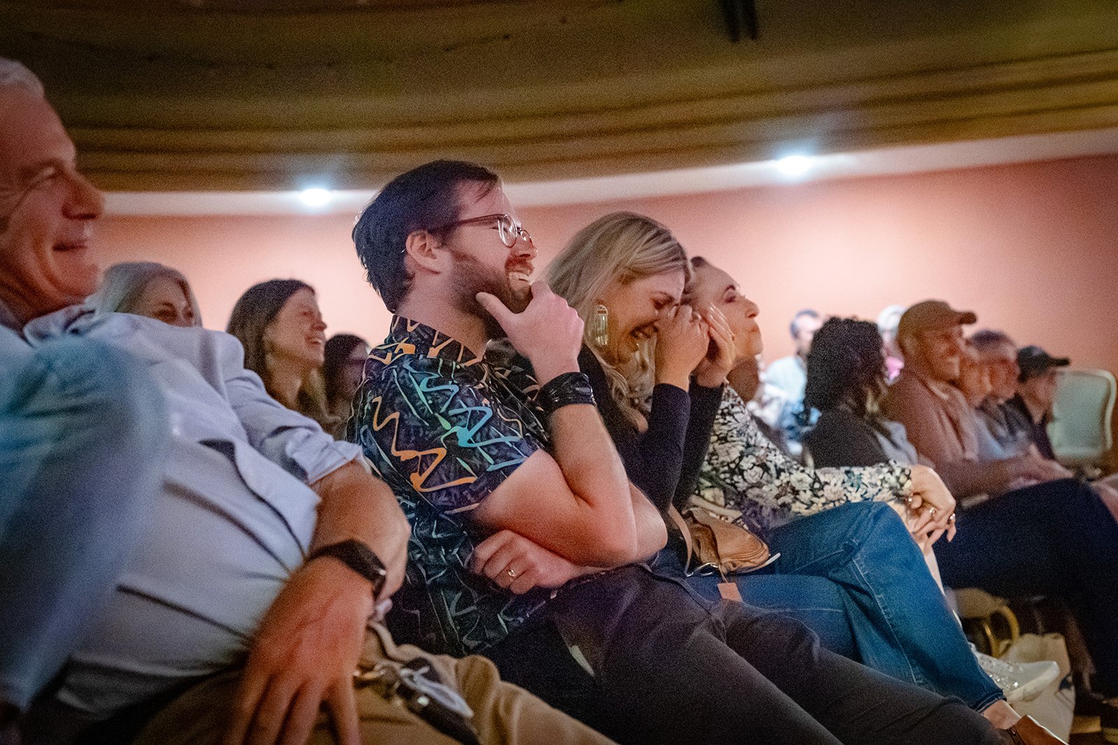 People sitting in an audience at a comedy or entertainment event, laughing and enjoying the moment. The room has warm lighting and a relaxed atmosphere.
