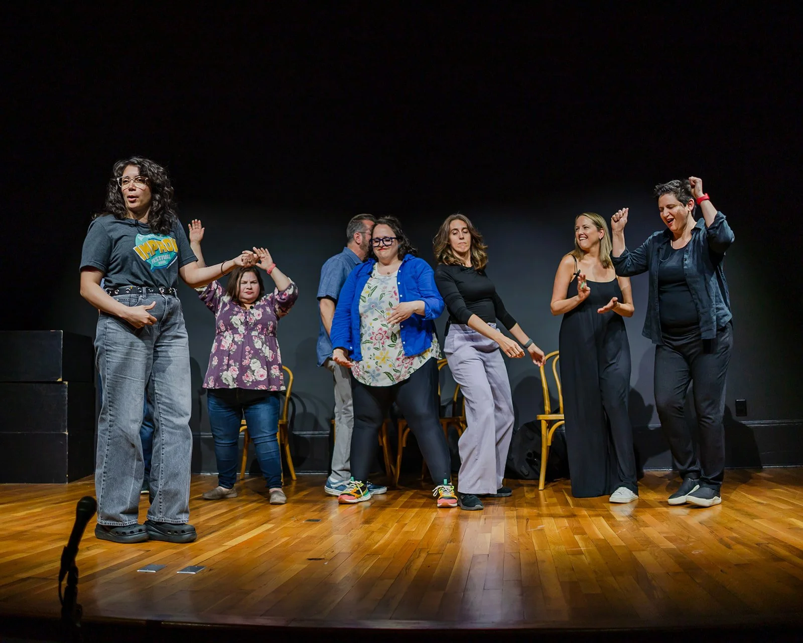 A group of seven people on stage participating in an improv comedy performance, some dancing and others watching.
