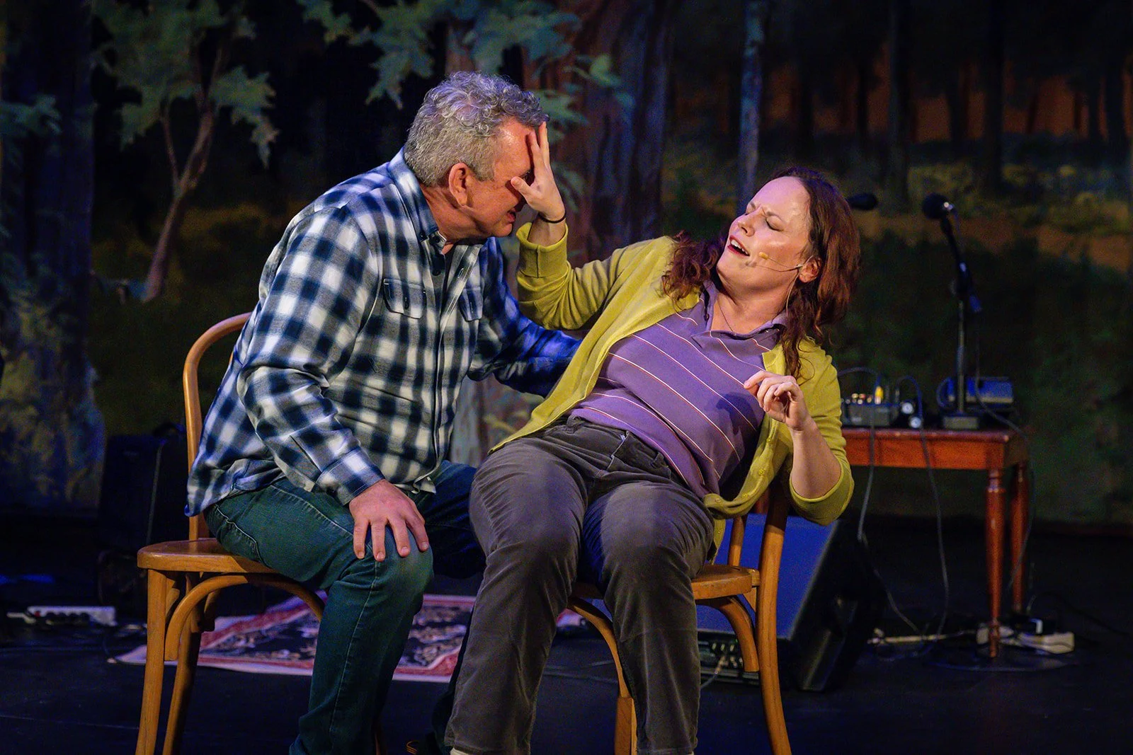 A theatrical performance showing two actors on stage, one man and one woman, engaging in a dramatic or comedic scene with expressive gestures and facial expressions. They are seated on wooden chairs, with a stage backdrop and sound equipment visible in the background.