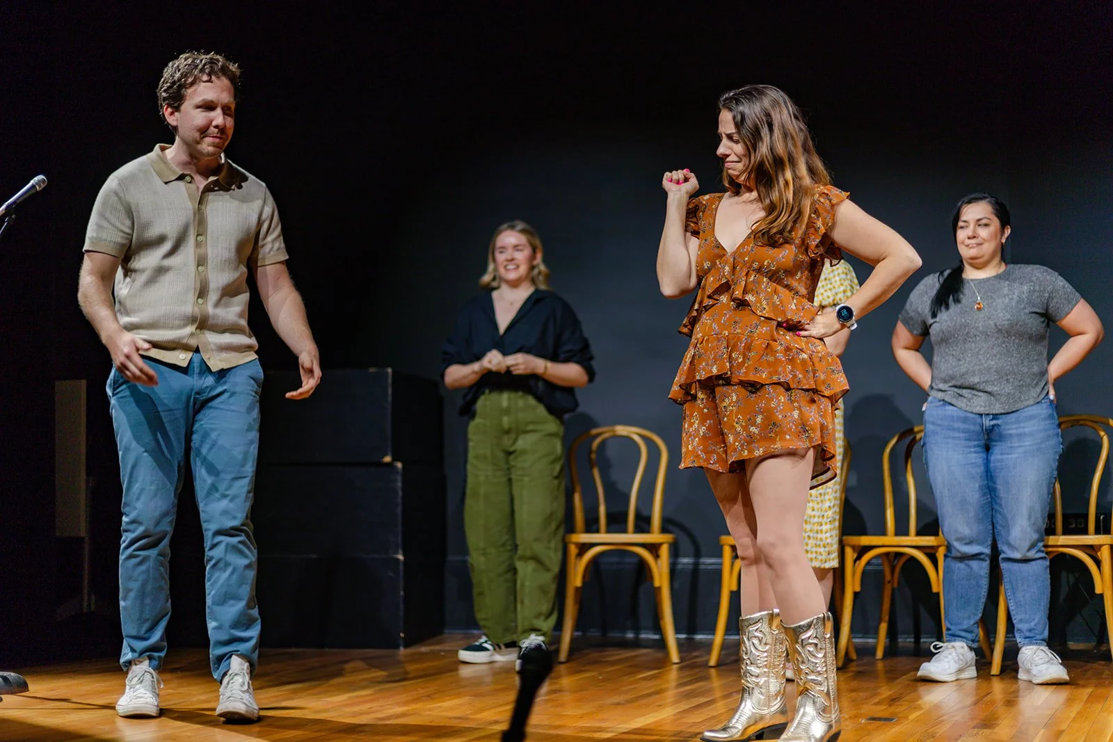 Group of women on stage, with one woman in a ruffled orange dress and silver boots emoting, while others watch and smile.