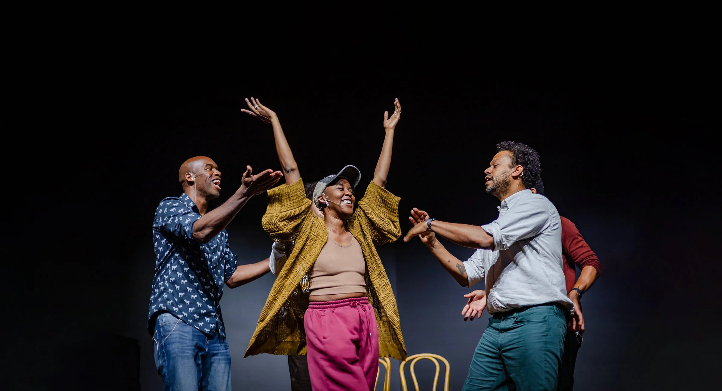 Group of five diverse people dancing and celebrating on stage, with three front and center, smiling and raising hands, dark background.