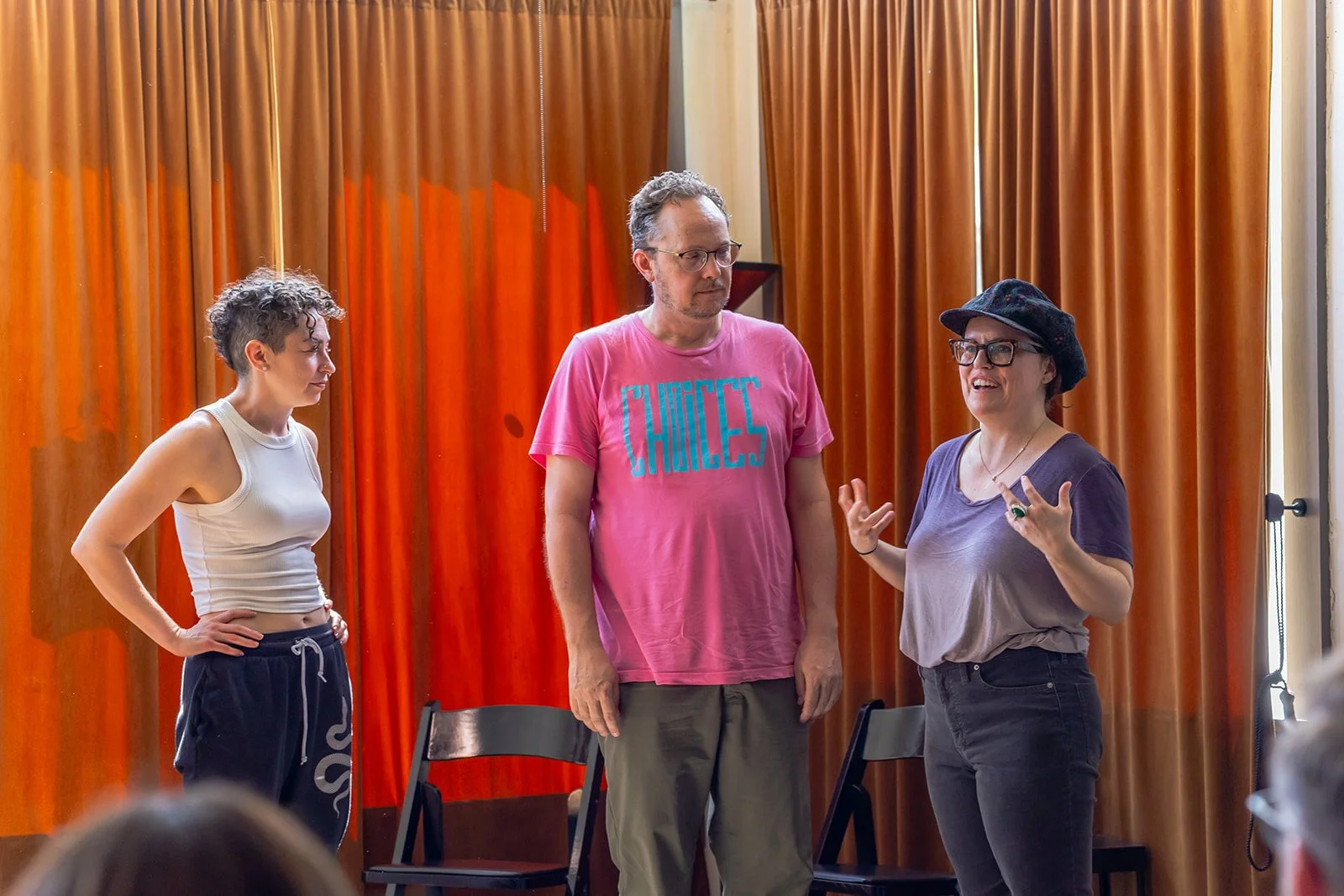 Three people standing and talking in a room with orange curtains, with chairs behind them and audience members partially visible in the foreground.