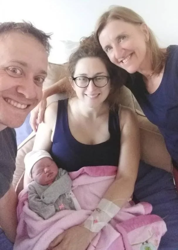 Newborn baby in mothers arms with her doula and partner standing next o her smiling
