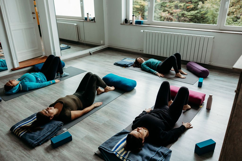 Group of five women practicing yoga in a bright, spacious room with large windows, lying on yoga mats with yoga blocks and water bottles nearby.