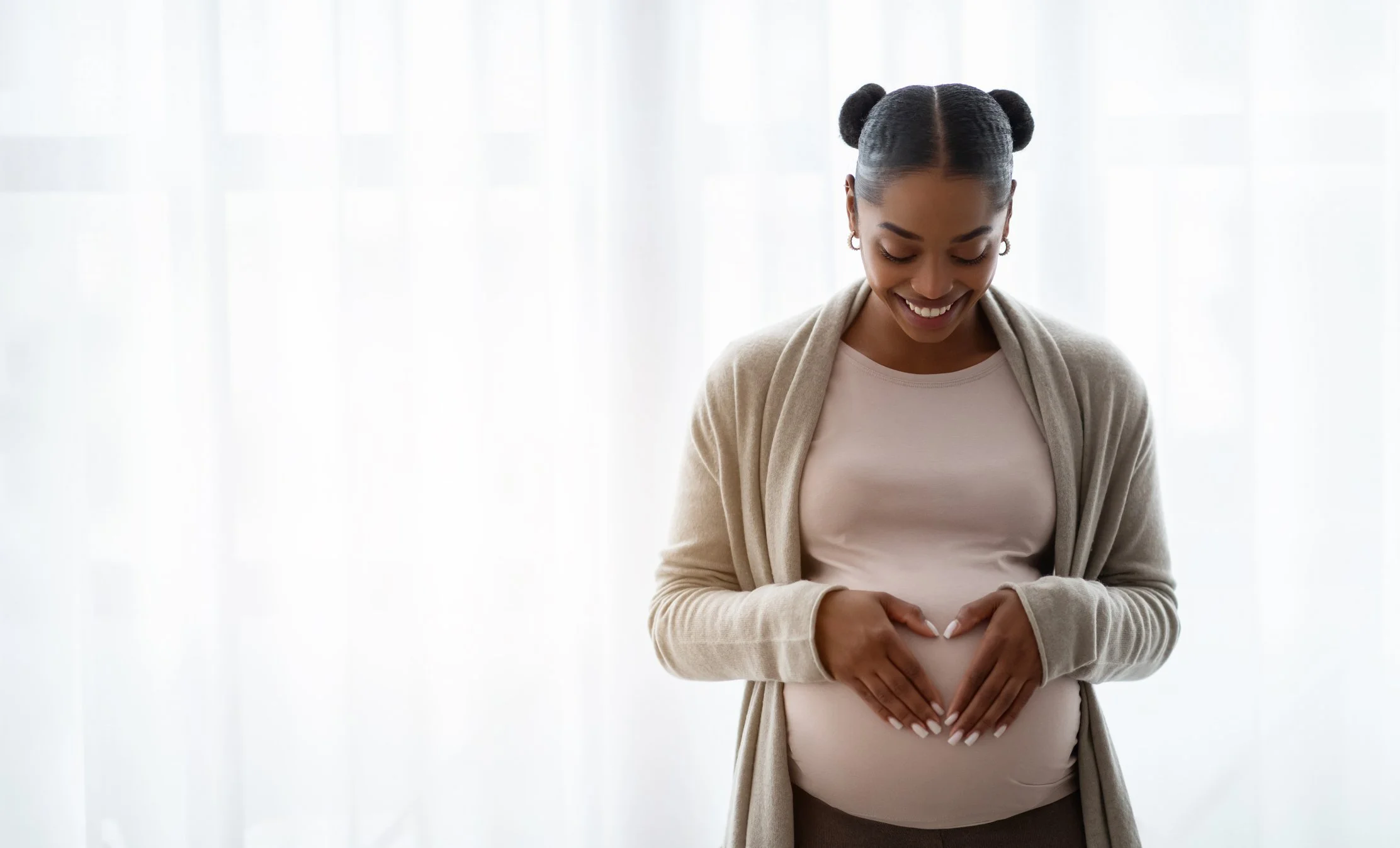 Pregnant woman smiling, hands forming heart on belly, standing near window.