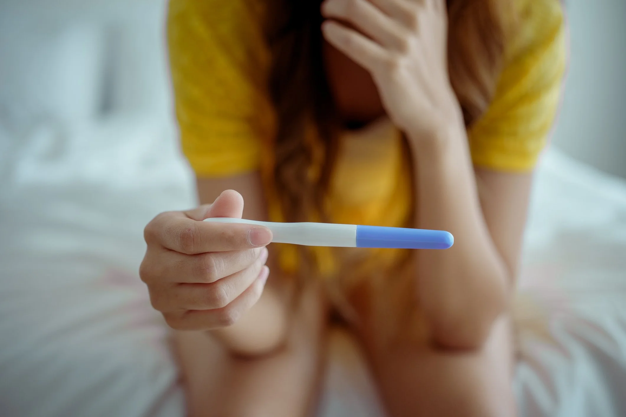 A woman in a yellow shirt holding a pregnancy test in her hand, sitting on a bed.