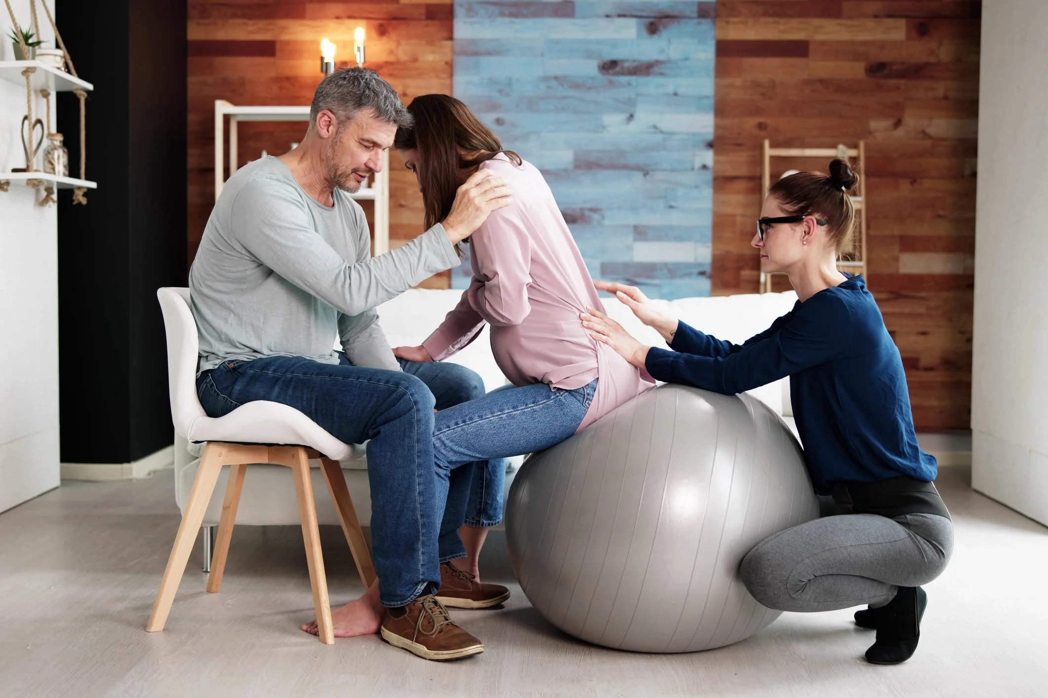 A pregnant woman is undergoing a physical exam with a healthcare professional using an exercise ball, while her partner and another woman provide support and comfort in a home setting.
